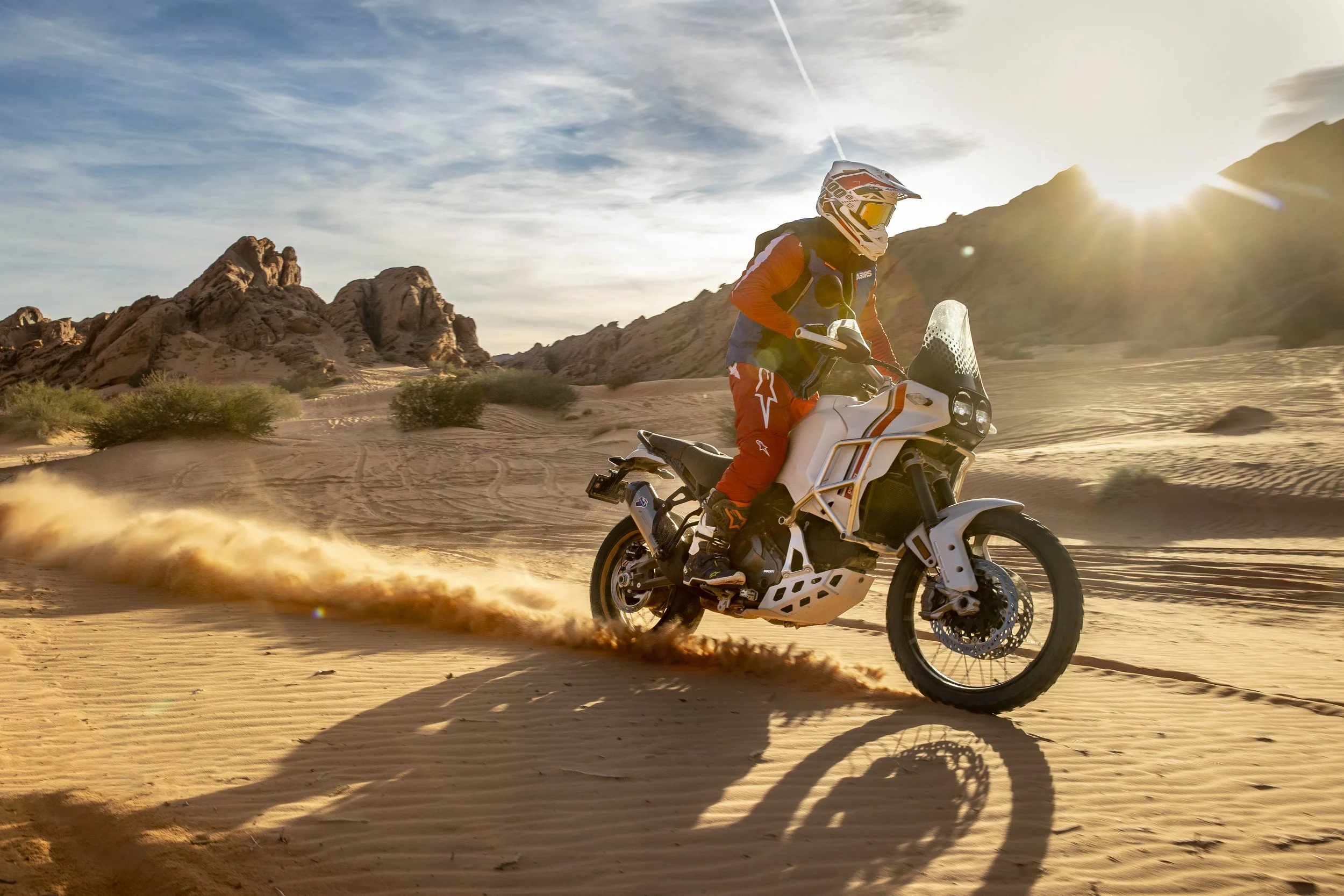 A person riding an adventure motorcycle on desert sand dunes during sunset with mountains in the background.