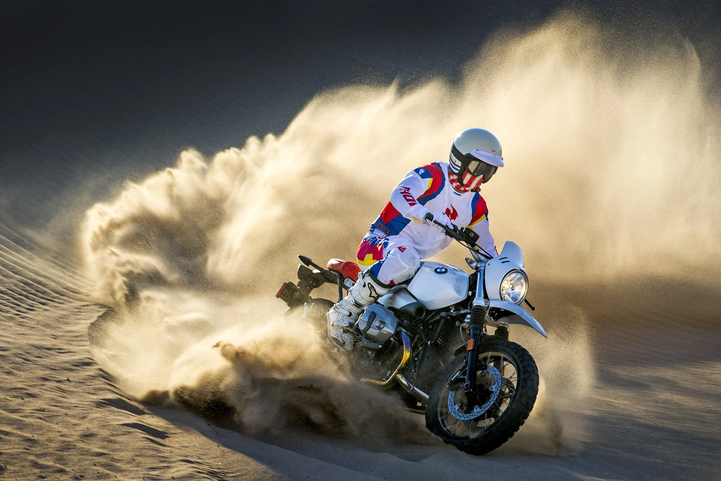 A motorcyclist in racing gear riding a BMW motorcycle at high speed on dirt, kicking up a trail of dust and sand during sunset.