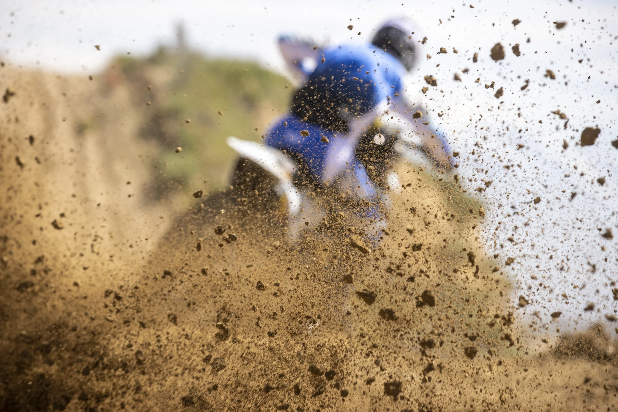 A person riding a dirt bike at high speed, kicking up a large cloud of dirt and rocks along a dirt trail, with a blurred background of trees and sky.