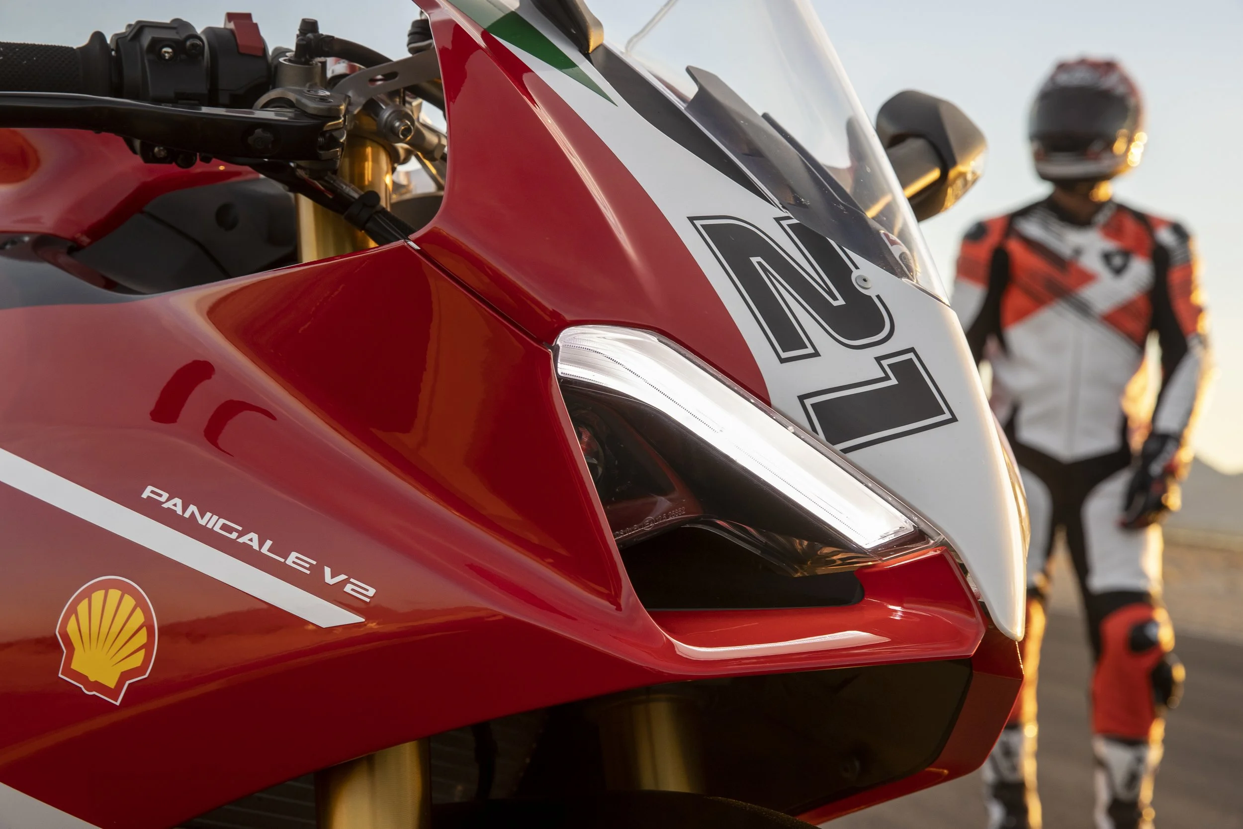Close-up of a red race motorcycle with a Honda logo, featuring a white number 1 and Shell sponsorship, with a blurred rider in a matching racing suit and helmet in the background.