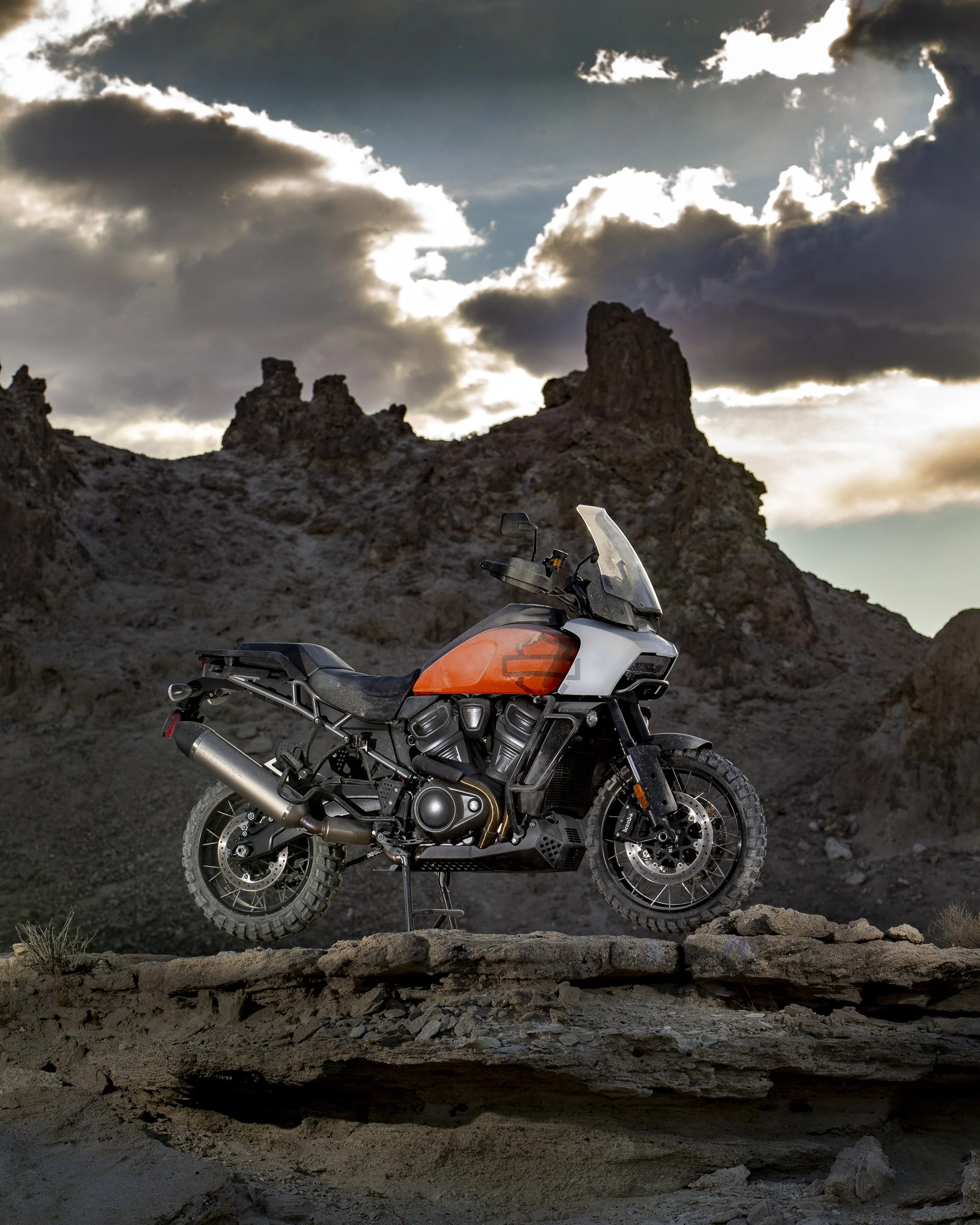 Adventure motorcycle parked on rocky terrain with rugged mountain rocks and cloudy sky in the background.