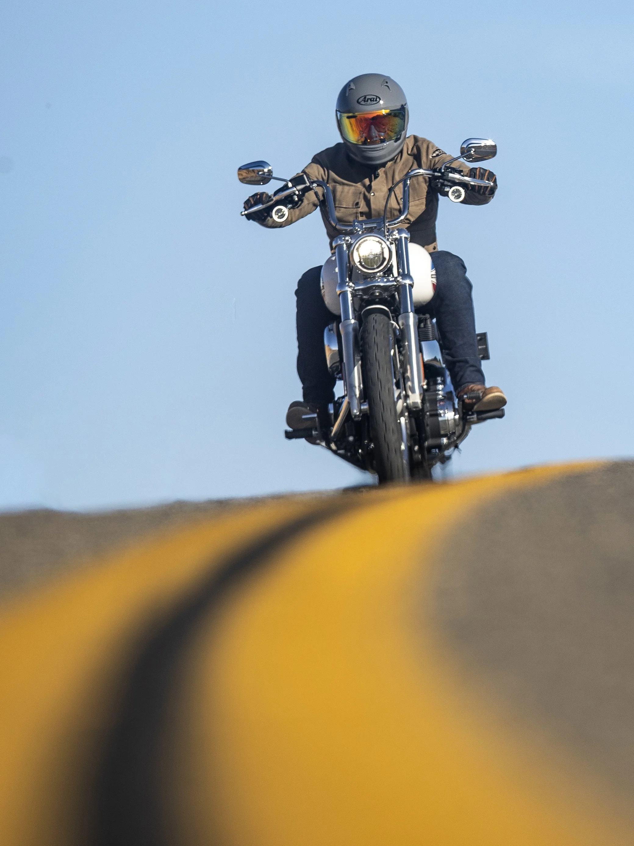 A person riding a motorcycle towards the camera, wearing a helmet and brown jacket, against a clear blue sky.