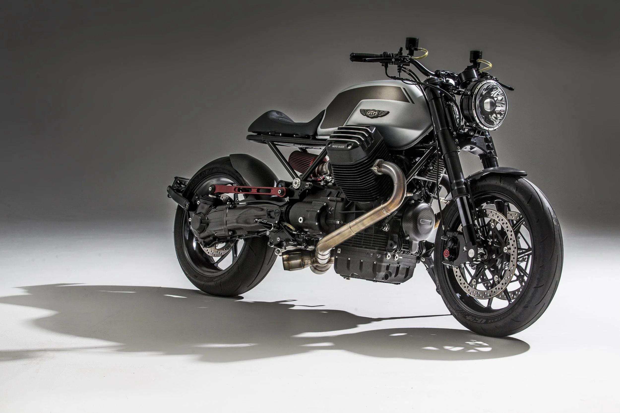 A custom black and silver motorcycle with a prominent round headlight, exposed engine components, black tires, and racing suspension, shot against a plain grey background.