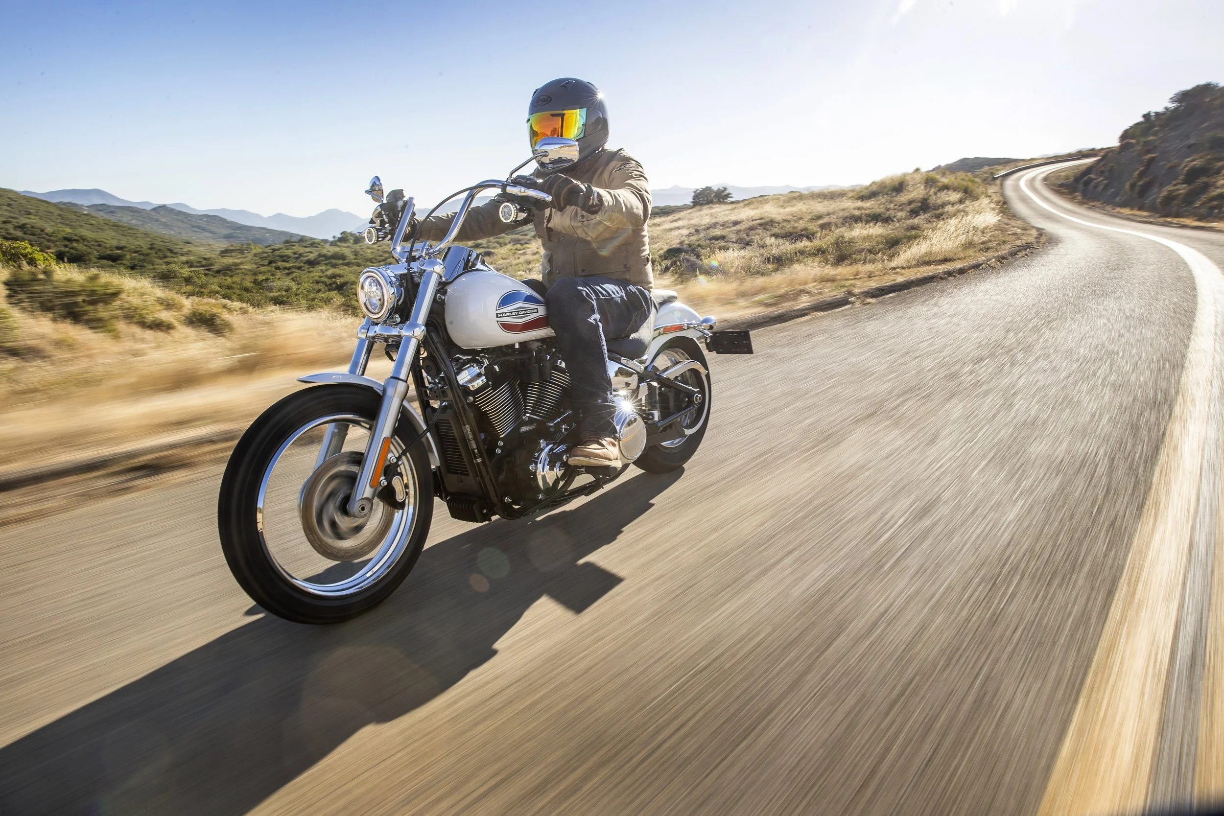 Person riding a Harley-Davidson motorcycle on a winding mountain road during daytime, wearing a helmet and jacket, with clear sky and scenic landscape in the background.
