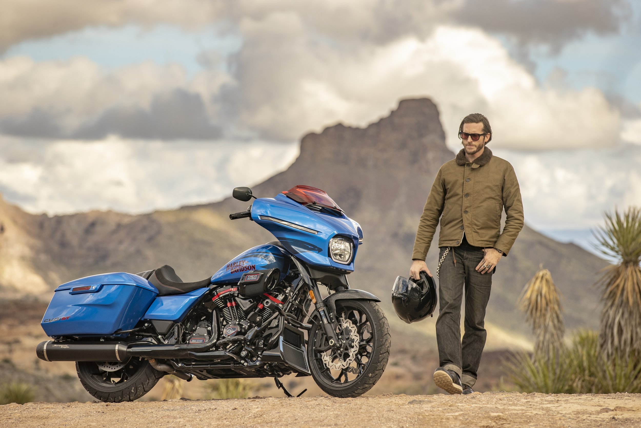 A man wearing sunglasses, a green jacket, and dark pants standing next to a blue Harley Davidson motorcycle in a desert landscape with mountains and cloudy sky in the background.