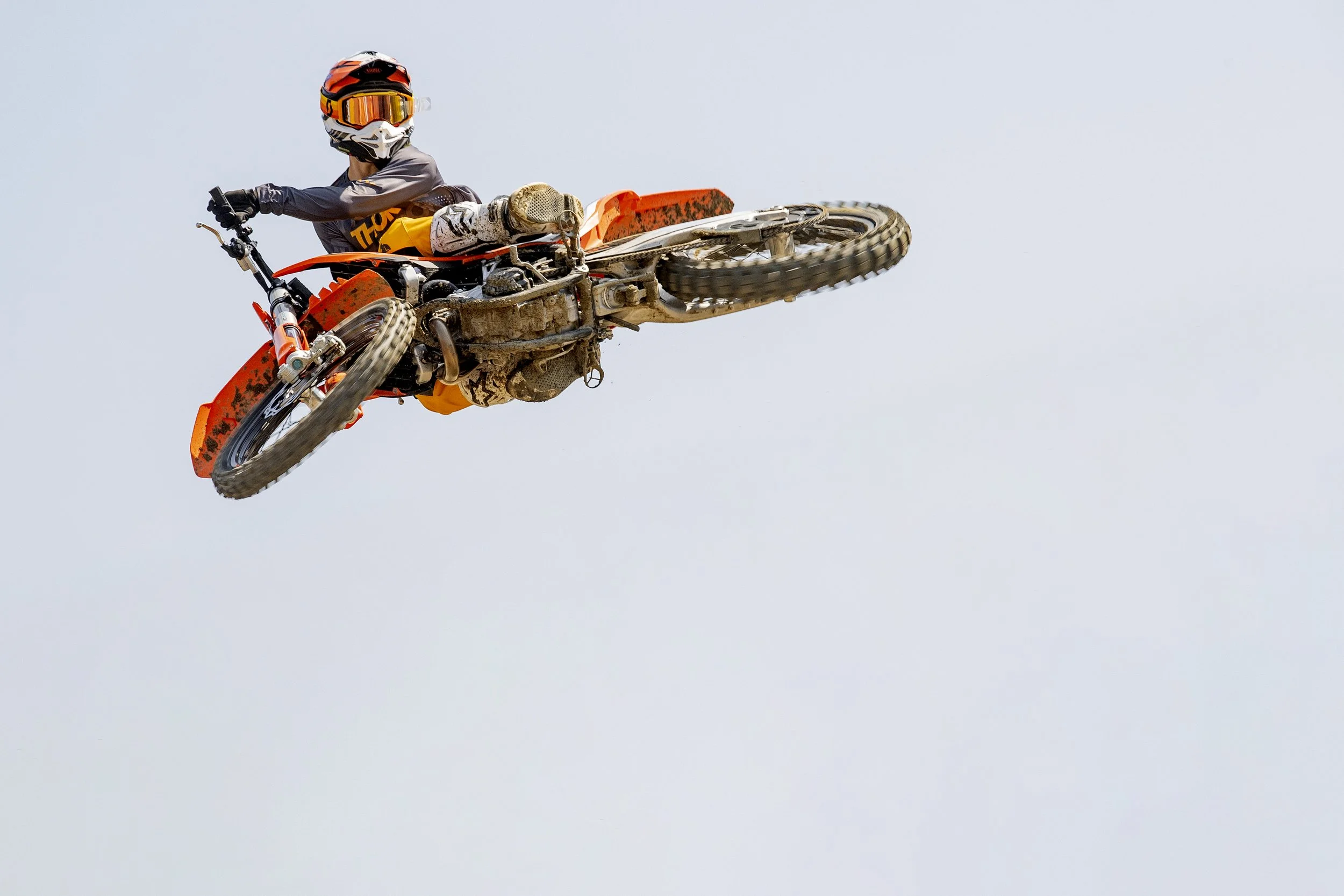 A motocross rider performing a mid-air stunt on an orange dirt bike, wearing a helmet and protective gear, against a clear sky background.