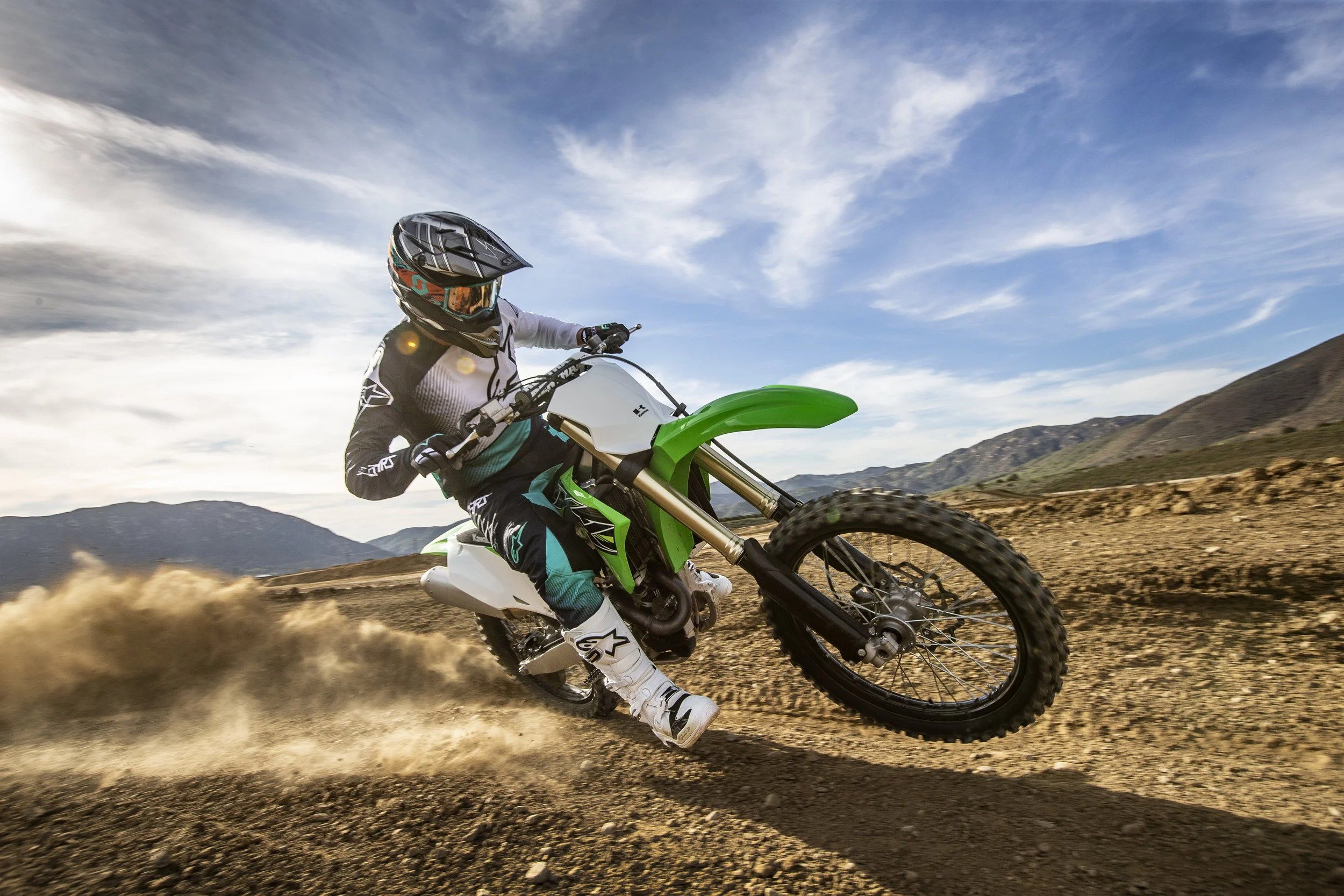 Motocross rider in full gear, riding a green and white dirt bike on a dirt trail with mountains and a partly cloudy sky in the background.