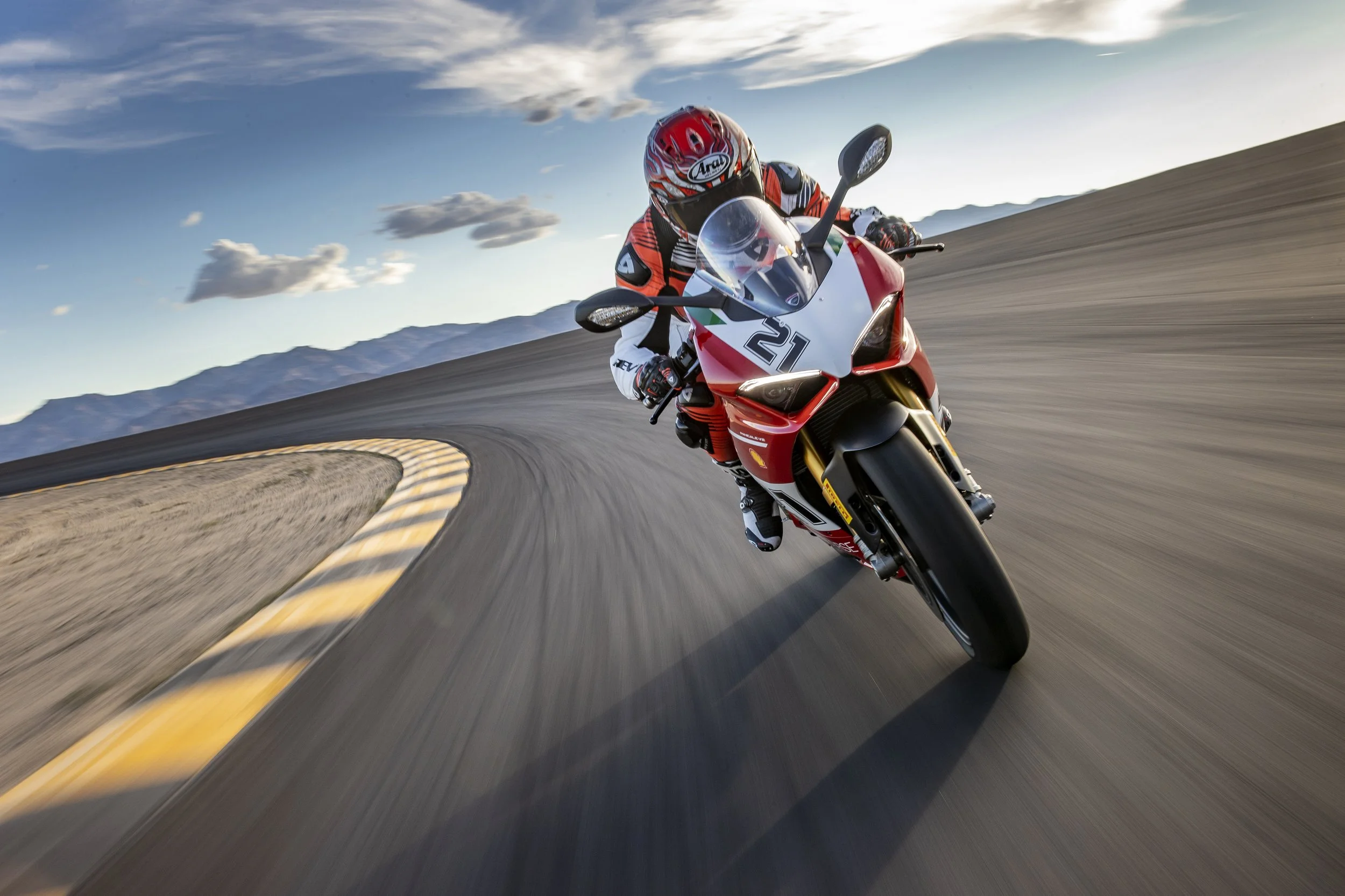 Motorcycle rider wearing red helmet and gear leaning into a turn on a racetrack with mountains and cloudy sky in the background.