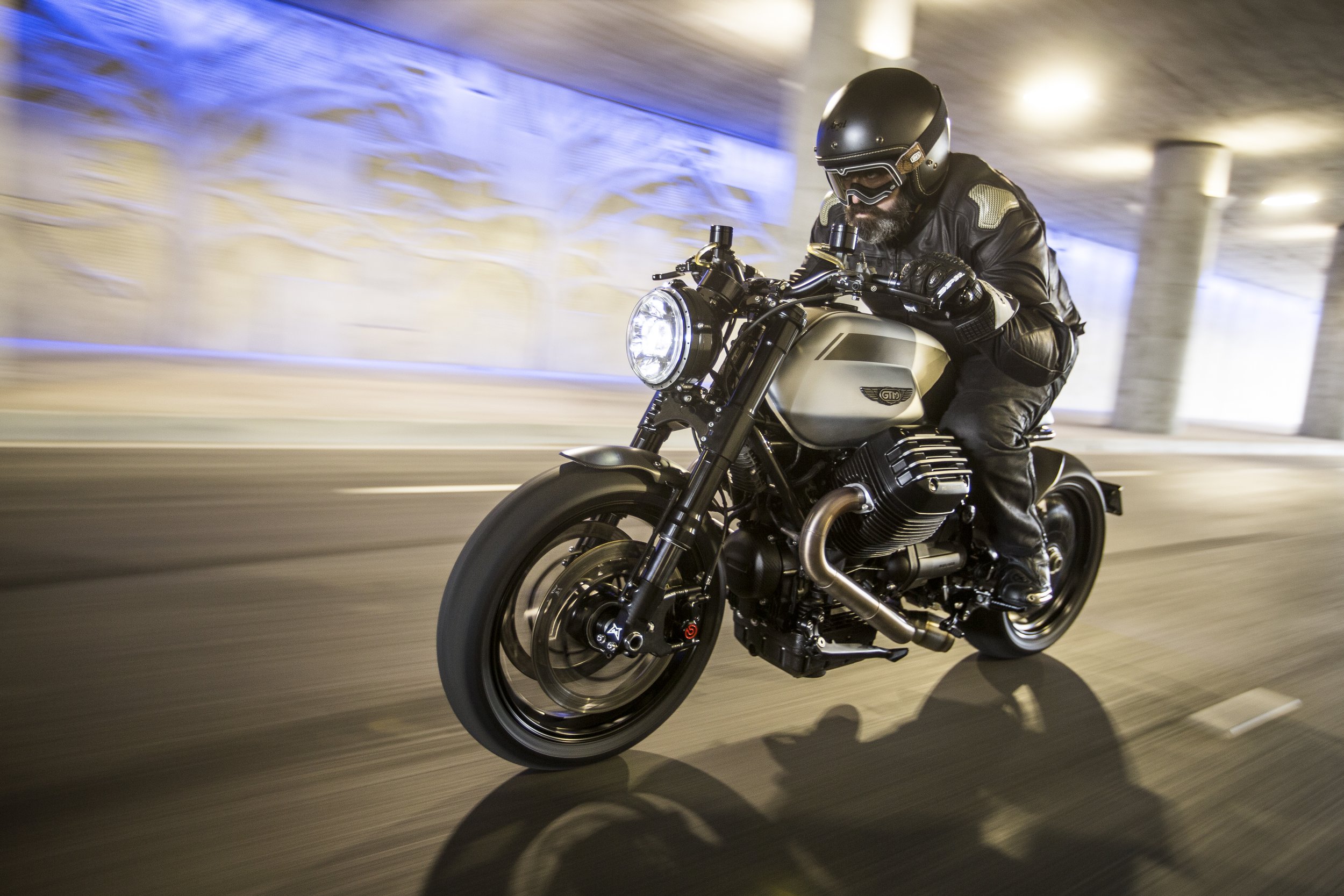 A man wearing a black helmet and leather jacket riding a silver motorcycle through an urban tunnel.