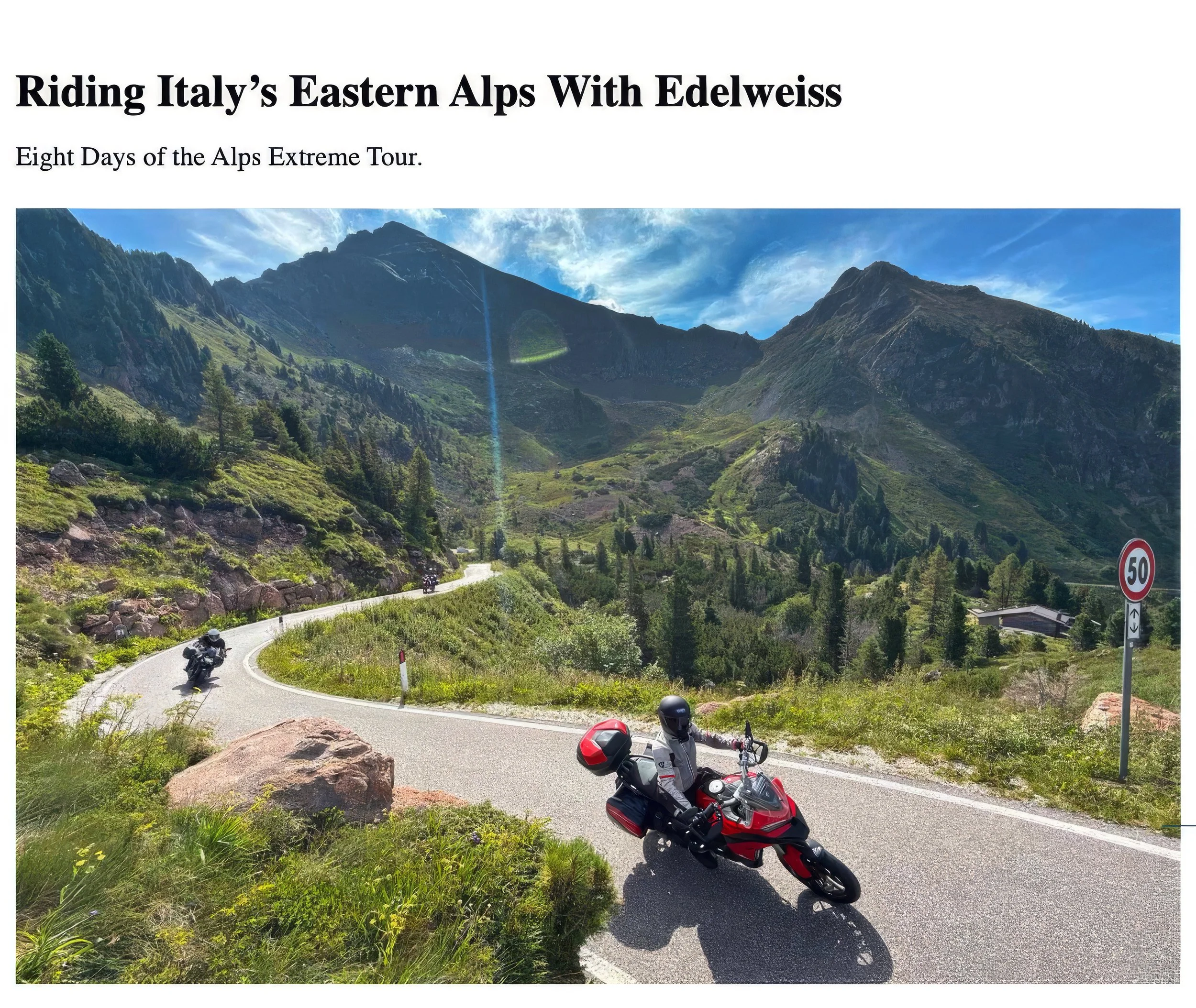 Motorcycles parked on a winding mountain road with green hills and rocky peaks in the background under a blue sky with clouds.