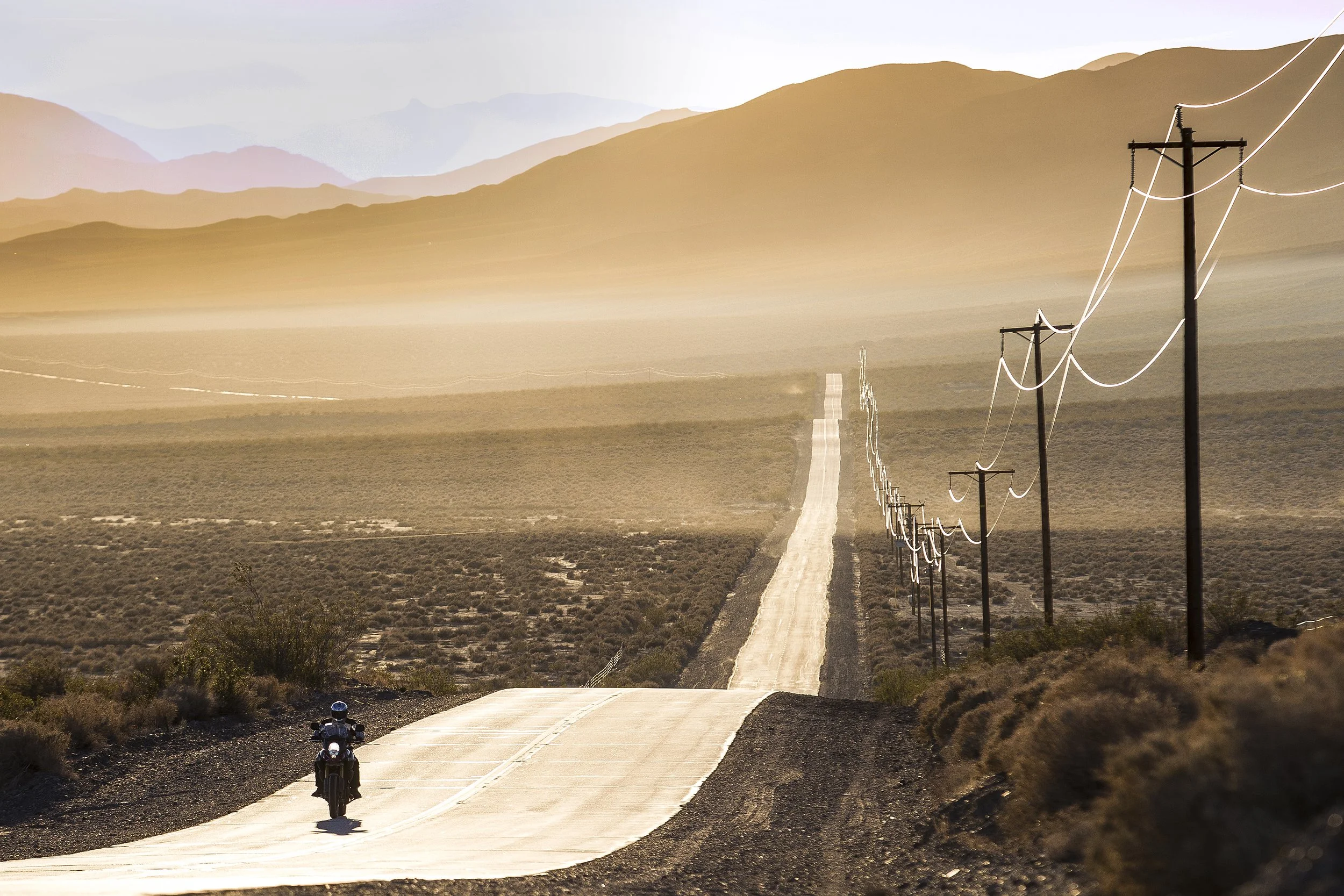 A motorcyclist riding on a long, straight desert road surrounded by arid desert landscape with sparse vegetation, power lines running alongside, and mountains in the distance under a clear sky.