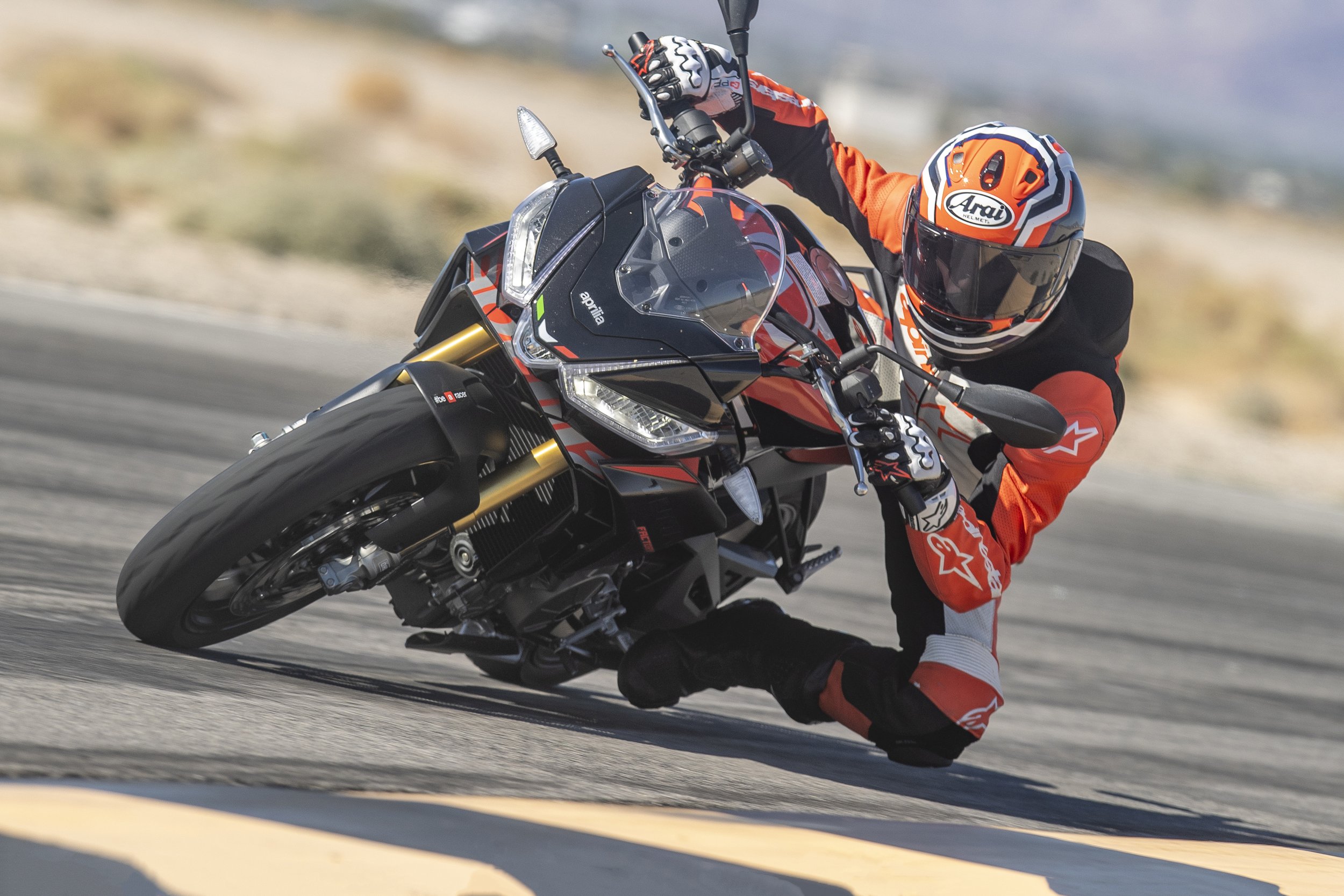 Motorcycle racer leaning sharply into a turn on the road, wearing orange and black racing gear, a helmet, and gloves, with the landscape blurred in the background.