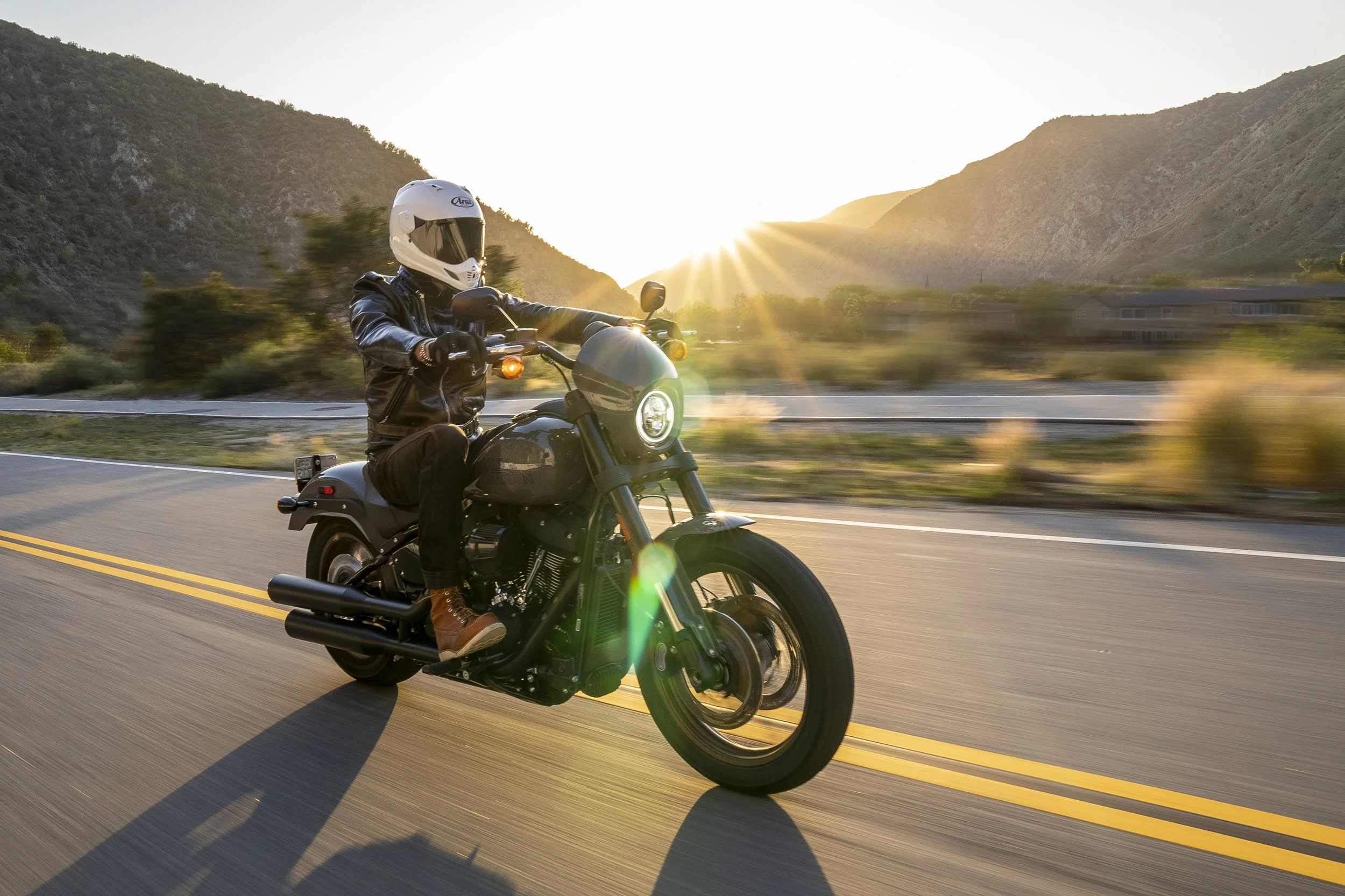Motorcyclist wearing a white helmet and black leather jacket riding a black motorcycle on a winding road during sunset with mountains in the background.