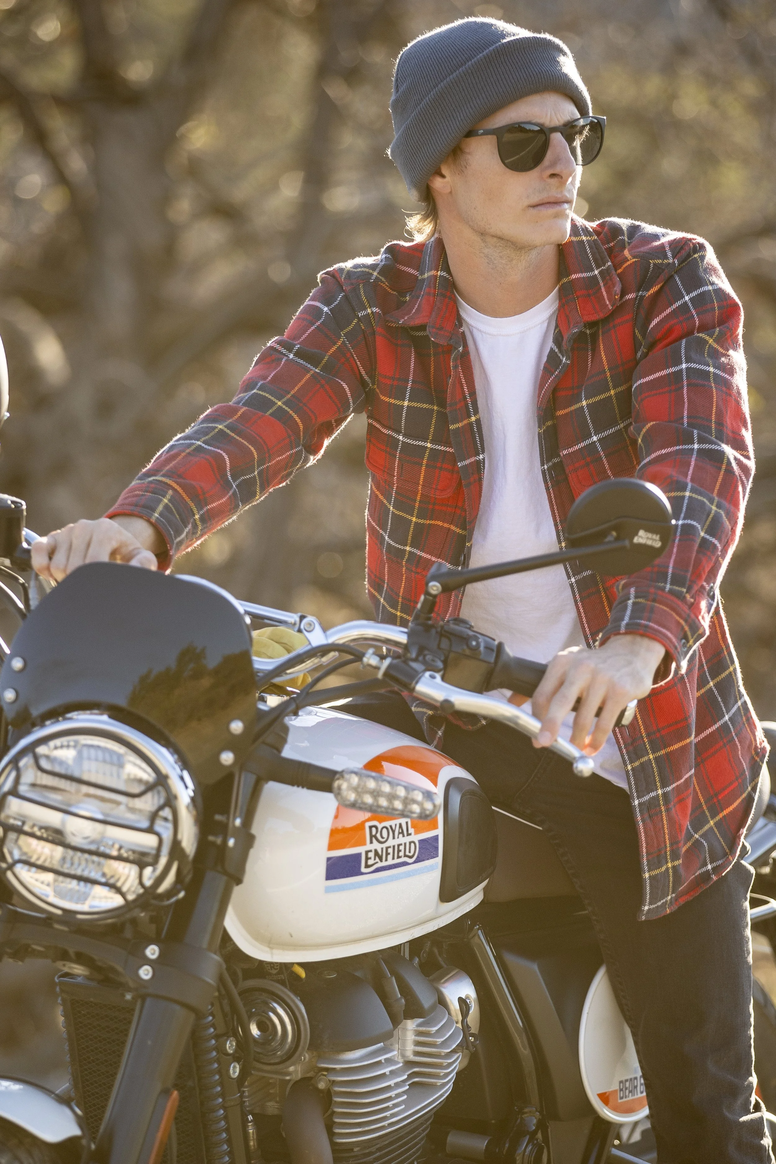 A man with blond hair, wearing a gray beanie, black sunglasses, a red plaid shirt, and black pants, sits on a Royal Enfield motorcycle outdoors during golden hour with trees in the blurred background.