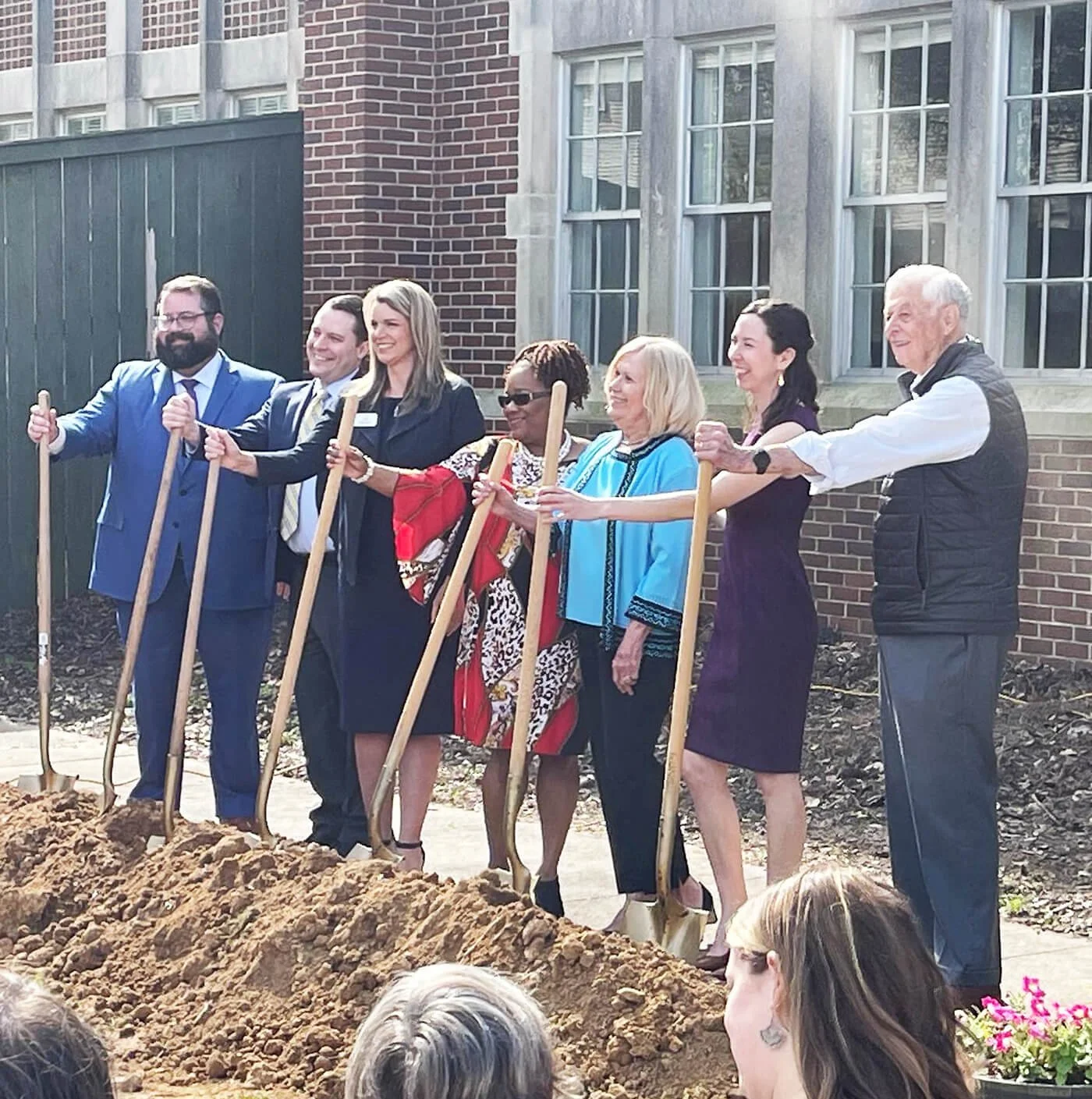 People participating in a groundbreaking ceremony, holding shovels with dirt in front, outdoors in front of a brick building.