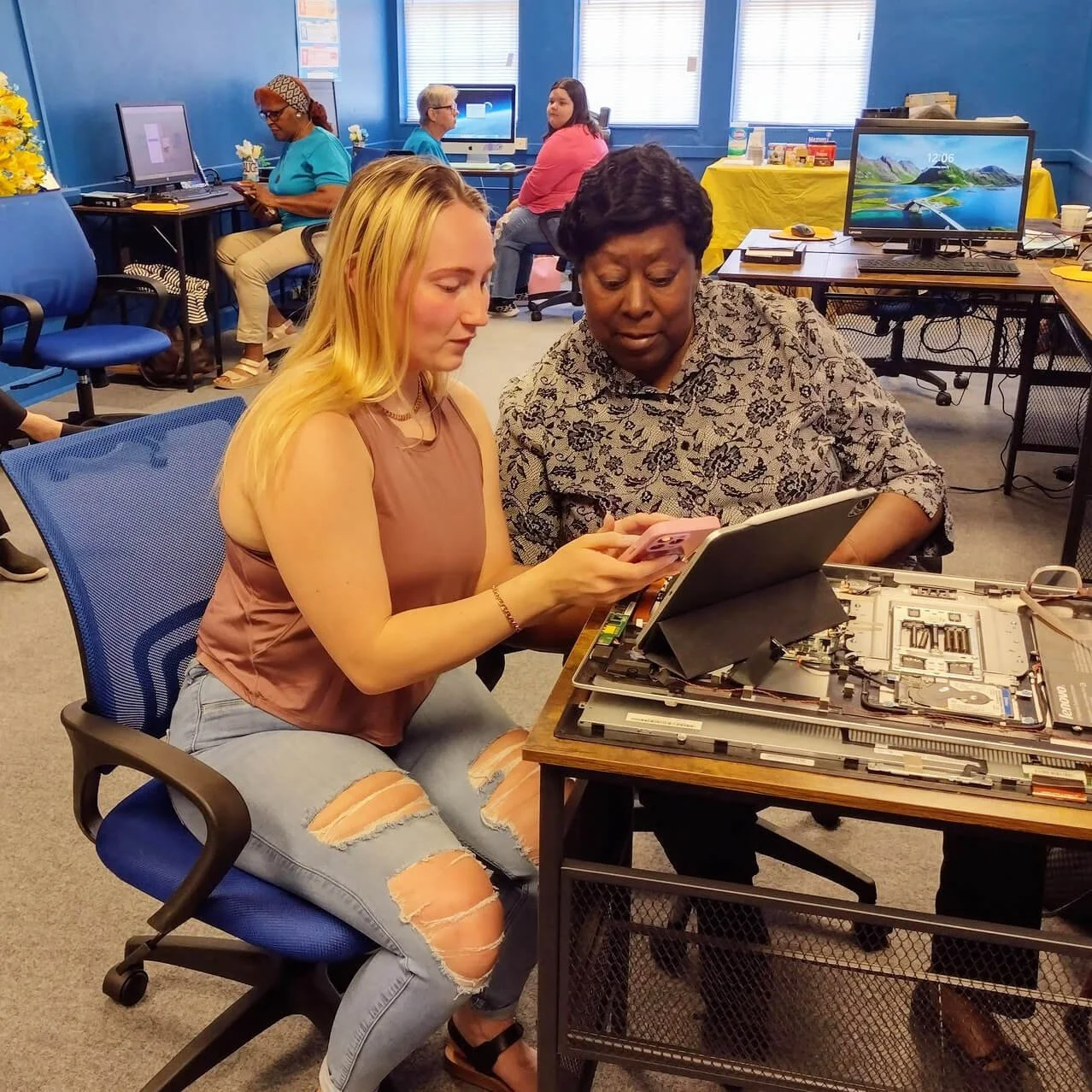 Two women working on a disassembled electronic device at a table, with several people working on computers in the background inside a room with blue walls and multiple computer monitors.