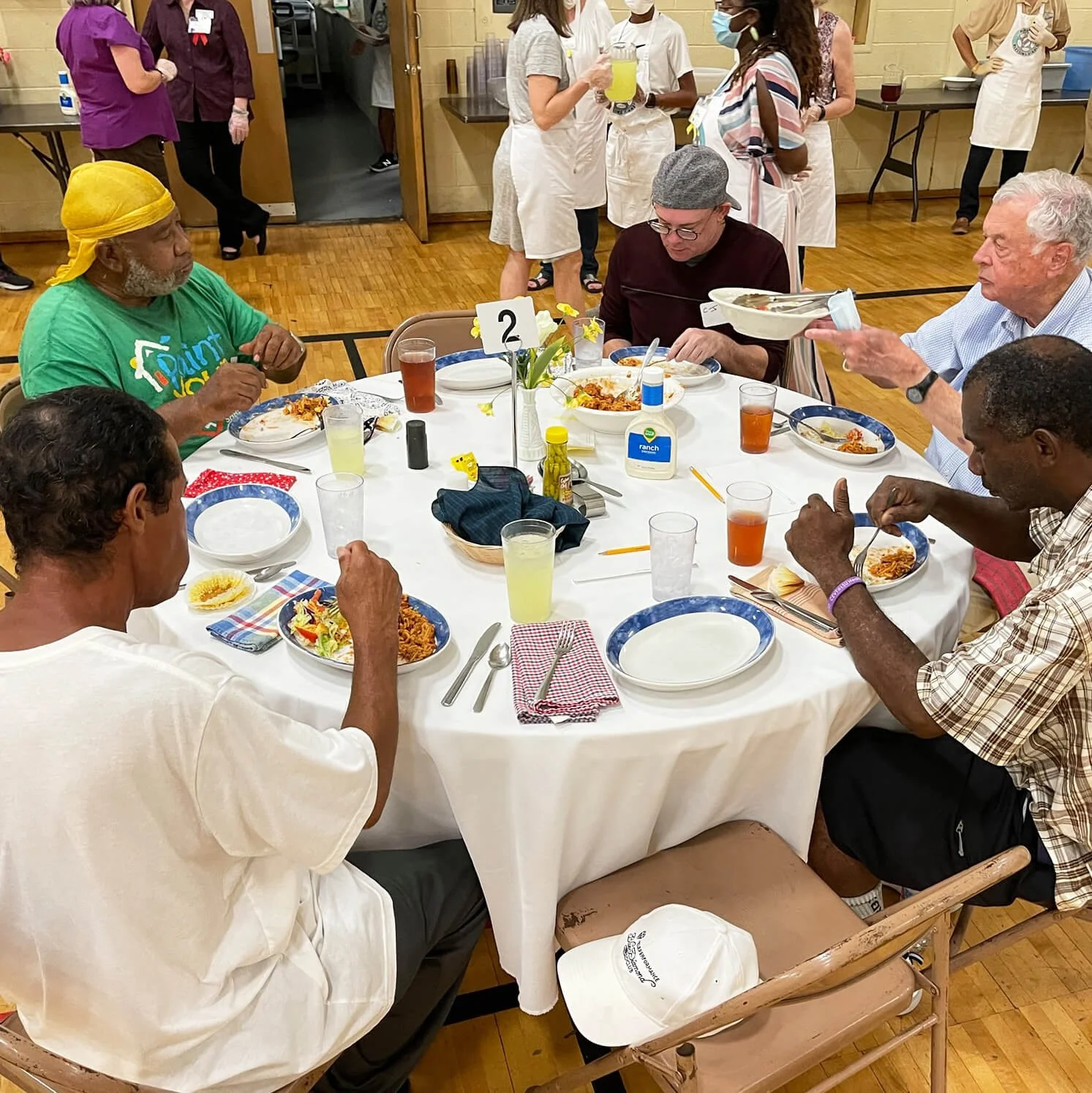 A group of people sitting around a round table enjoying a meal. The table is set with plates of food, drinks, napkins, utensils, and condiments. People are eating, and some are serving or pouring drinks. The setting appears to be a communal dining event with others in the background.