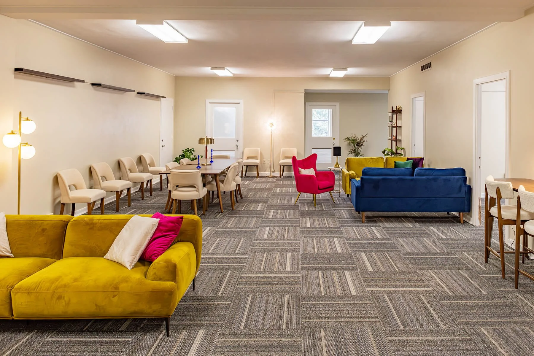 A spacious, brightly lit lounge area with colorful furniture, including yellow, red, blue, and green sofas, along with beige chairs and a large dining table. The room features modern decor, floor lamps, and plants, with windows and doors providing natural light.