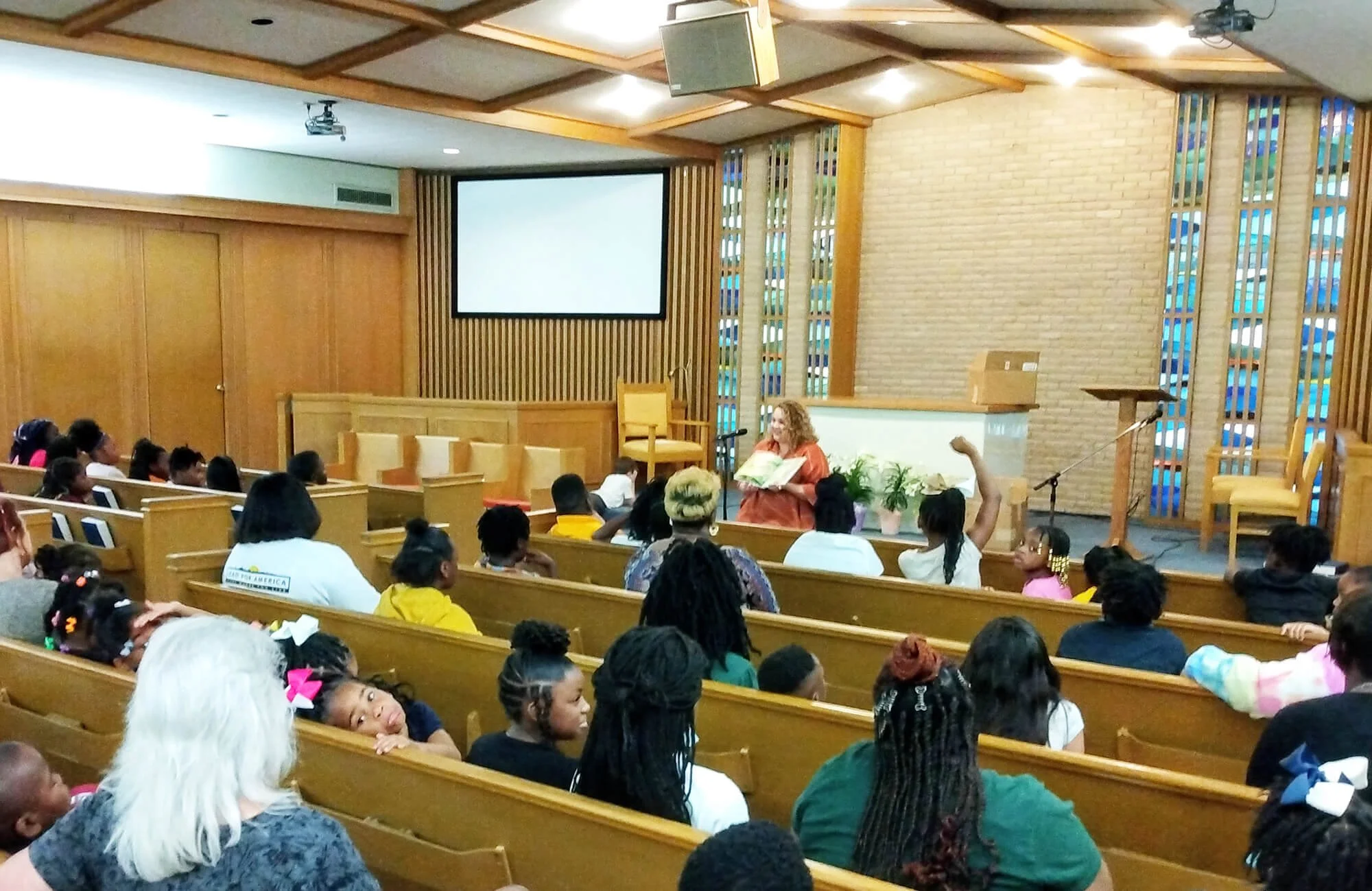 Inside a chapel with wooden walls and seating, a diverse group of children and adults seated facing a woman standing at the front, holding an open book. There is a large blank screen above and to the left of the woman, with a microphone stand nearby.