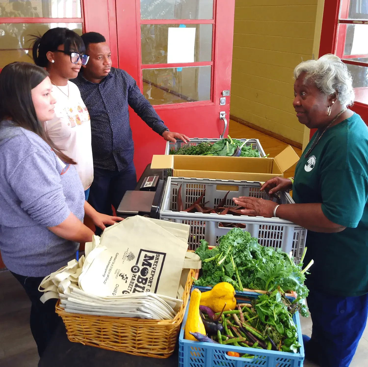 A diverse group of people standing at a produce stand, talking to an older woman behind the table, which displays fresh vegetables. There are tote bags on the table, and the setting appears to be indoors with red and yellow walls.