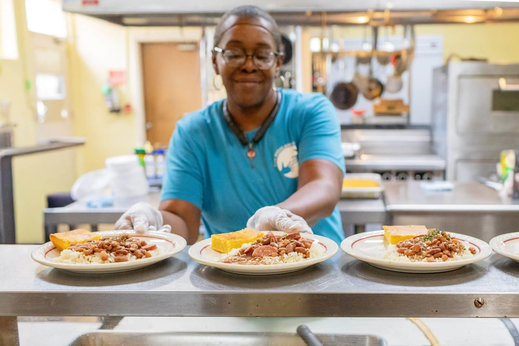 A woman wearing glasses, a blue shirt, and gloves is serving plates of food in a cafeteria setting. The plates contain rice, beans, and a piece of cornbread. The background includes kitchen appliances and shelves.