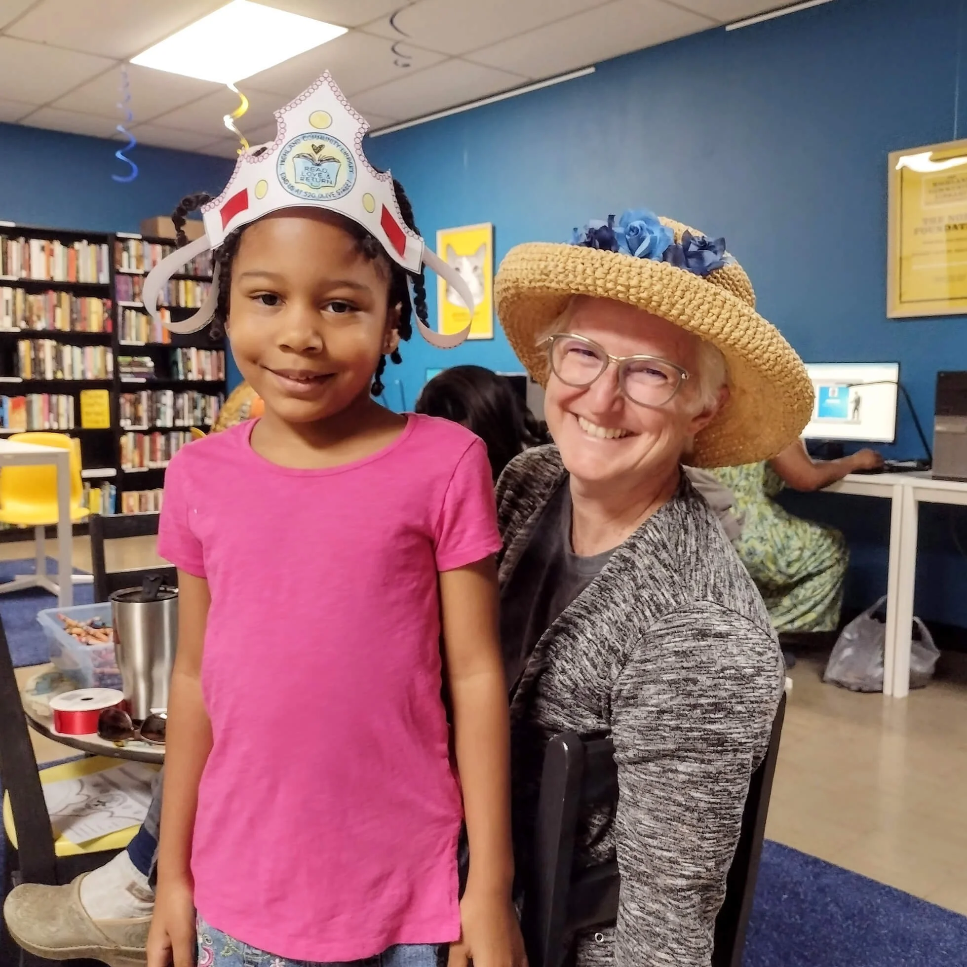 A young girl in a pink shirt and a woman wearing a straw hat with blue flowers, both smiling. The girl is wearing a paper crown and they are in a library with bookshelves in the background.