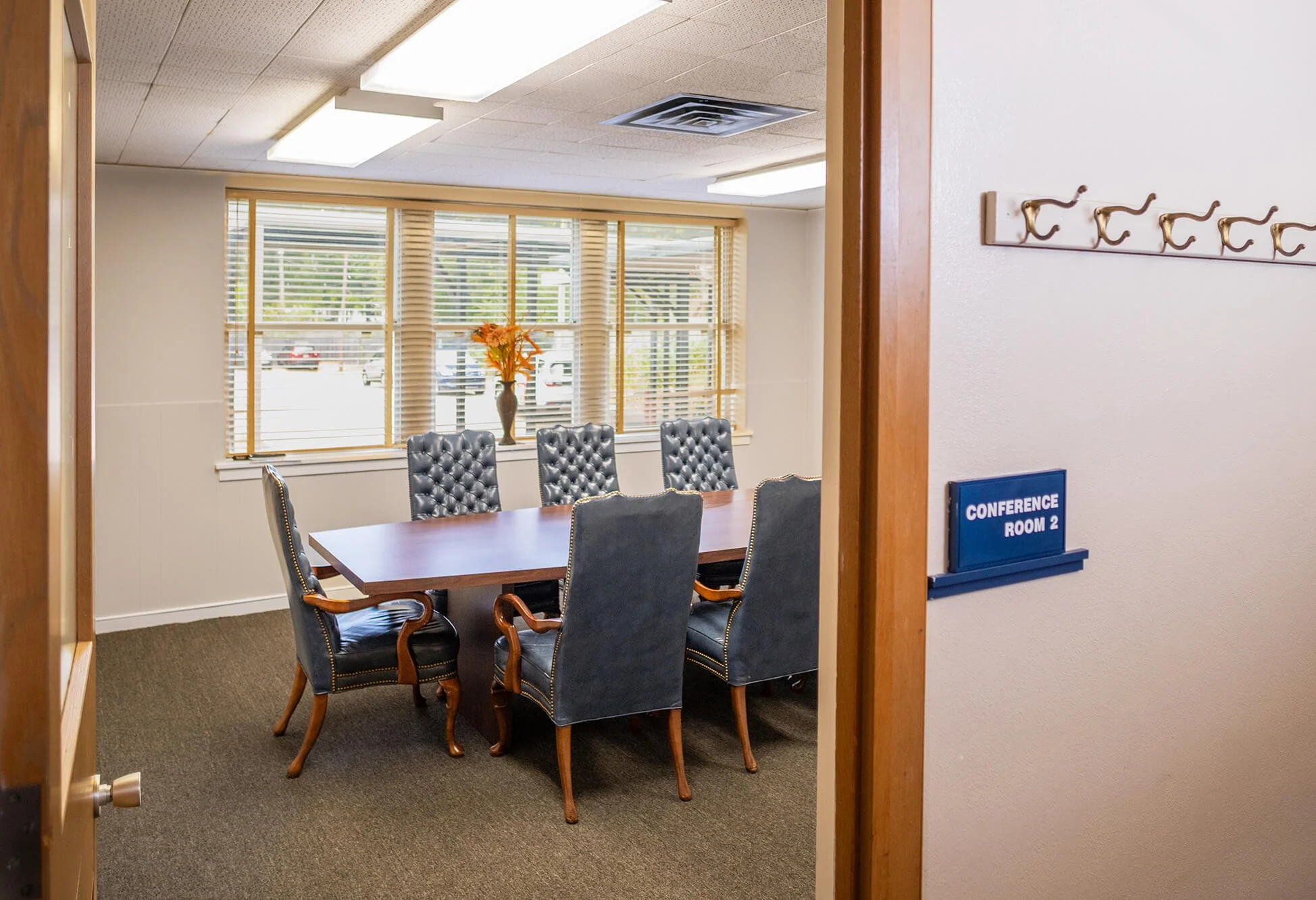 A small conference room with a rectangular wooden table surrounded by six high-backed, tufted leather chairs. The room has large windows with blinds, a decorative vase with orange flowers on the windowsill, and is illuminated by ceiling lights. There is a blue sign on the wall that reads 'Conference Room 2,' and a coat rack mounted on the wall.