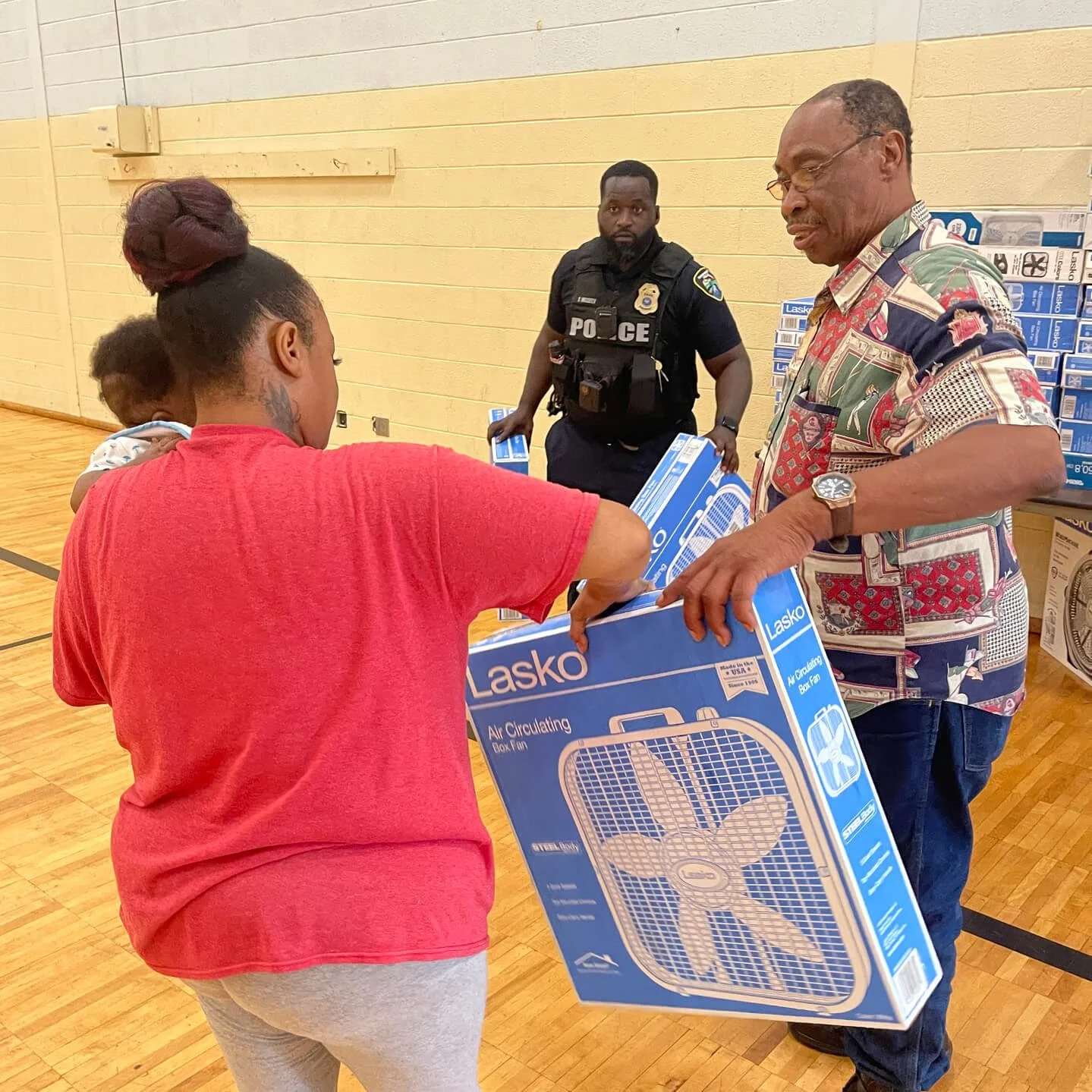 A woman in a red shirt hands a box fan to a man wearing a colorful shirt. A police officer stands in the background observing.