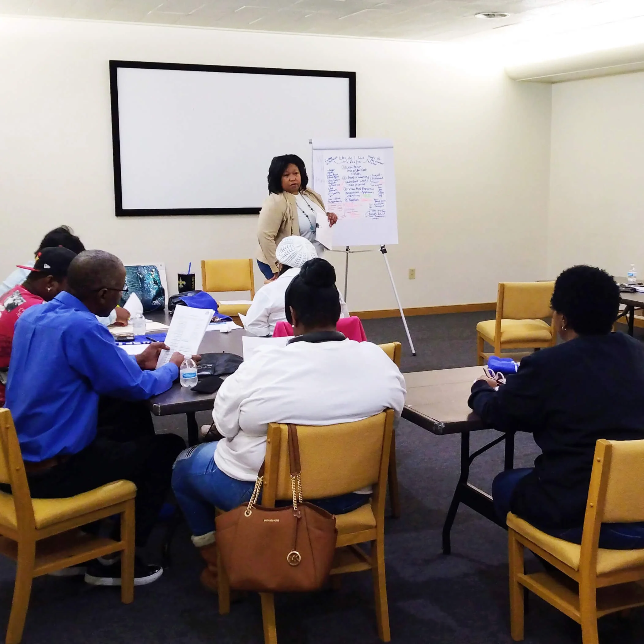 A woman standing and speaking at the front of a room during a presentation or workshop, with a flip chart on an easel and a large white projection screen behind her, while an audience of people sit at tables listening.