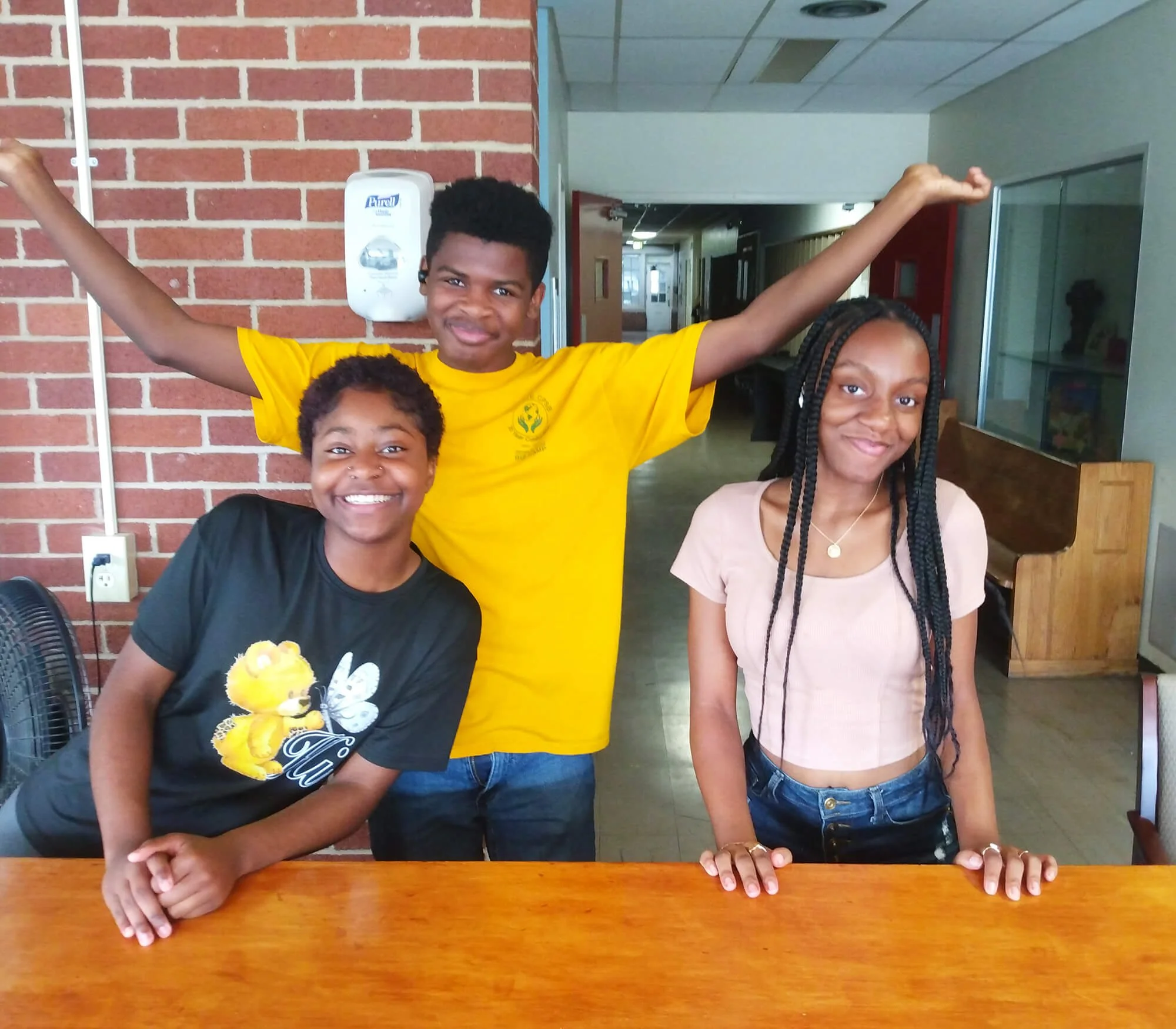 A group of three young people posing inside, all smiling at the camera. The setting appears to be an indoor space with brick, glass, and beige walls.
