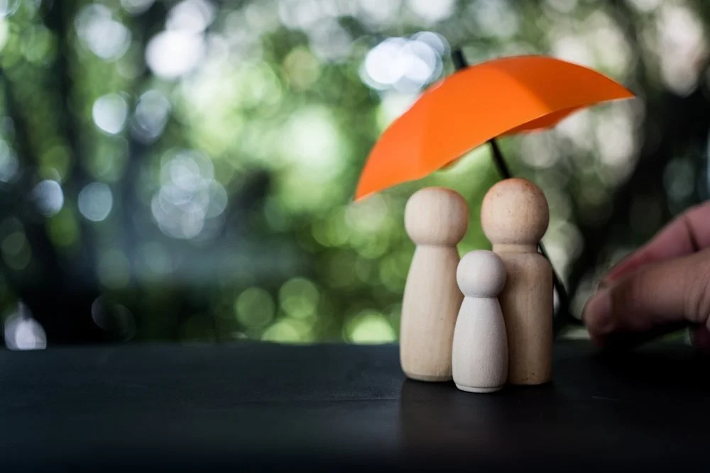 Three wooden figurines of different sizes with an orange umbrella above them, with a blurred green outdoor background.