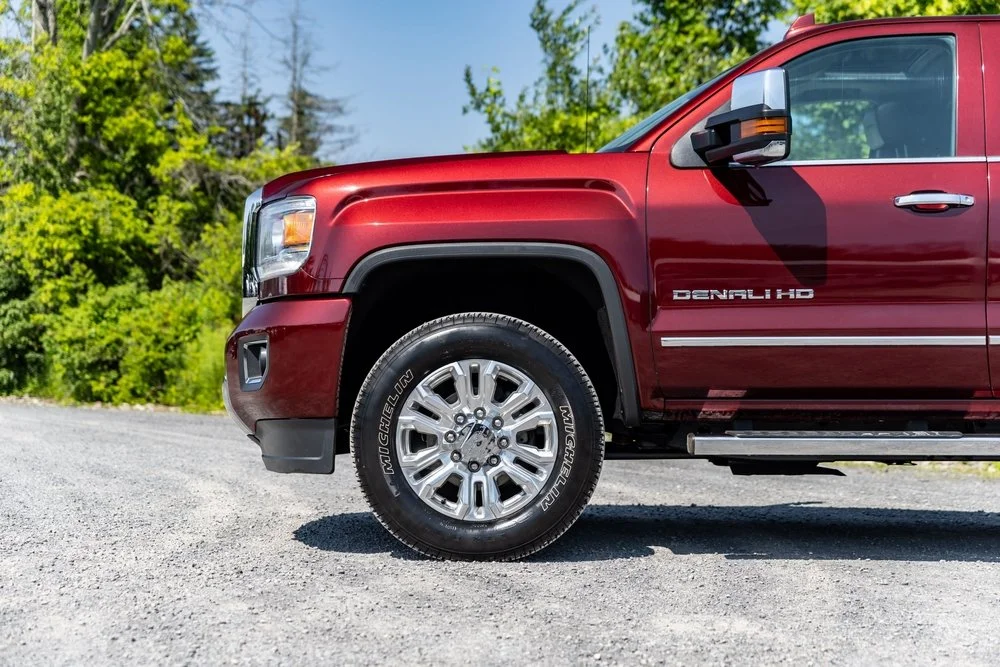 Close-up of front part of a red GMC Denali HD pickup truck on gravel road with green trees in background.