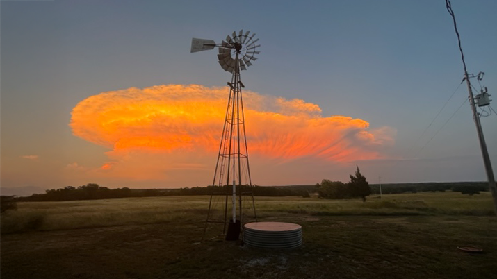A windmill standing in a grassy field during sunset, with a large orange cloud and a utility pole with power lines in the background.