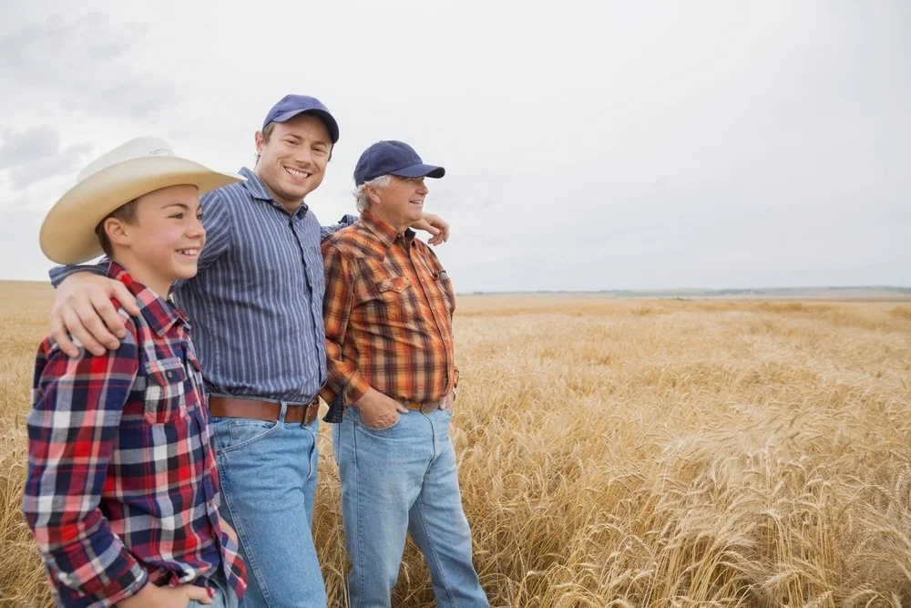 Three people smiling and looking into the distance while standing in a wheat field, two adults and a child, all wearing plaid shirts and hats.