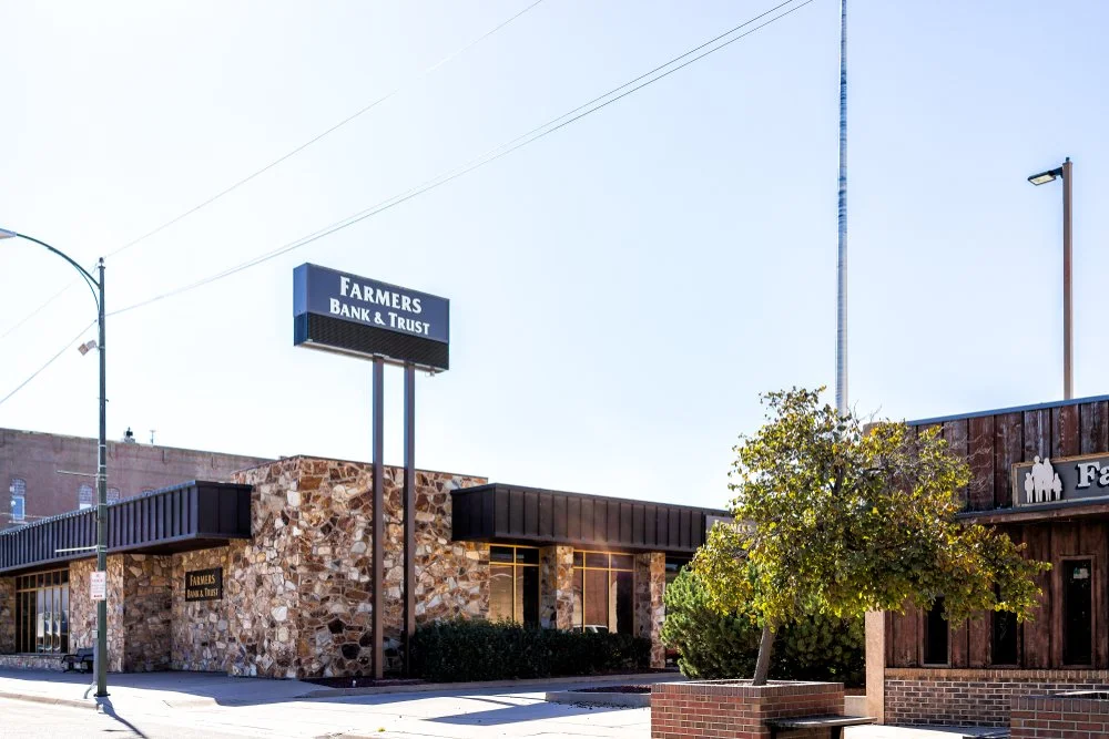 Exterior of a bank building with a sign that reads "Farmers Bank & Trust" and a stone facade, with neighboring wooden building, trees, and streetlamp.