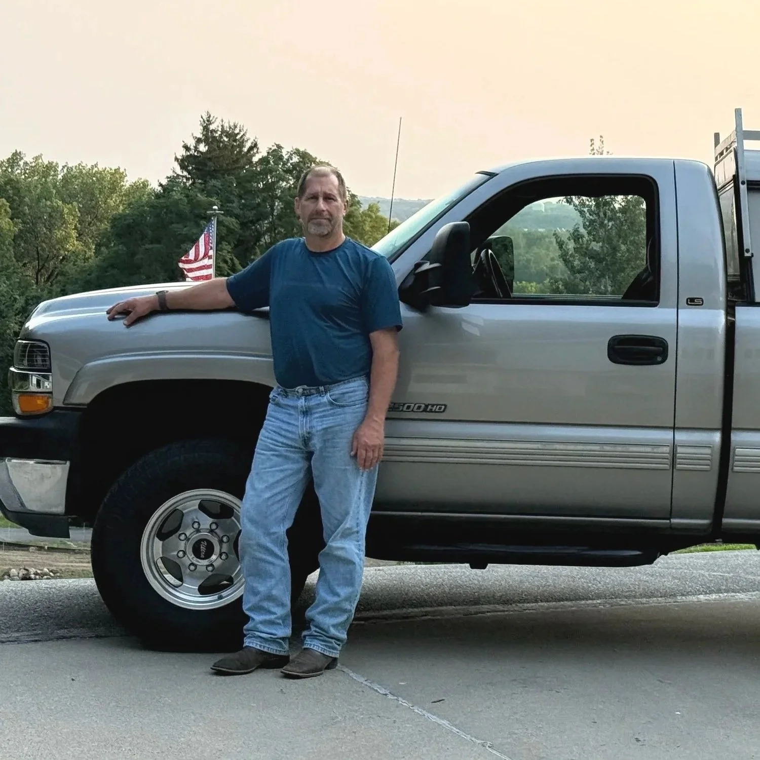 Daryl standing next to his silver pickup truck, outdoors with trees and an American flag in the background.