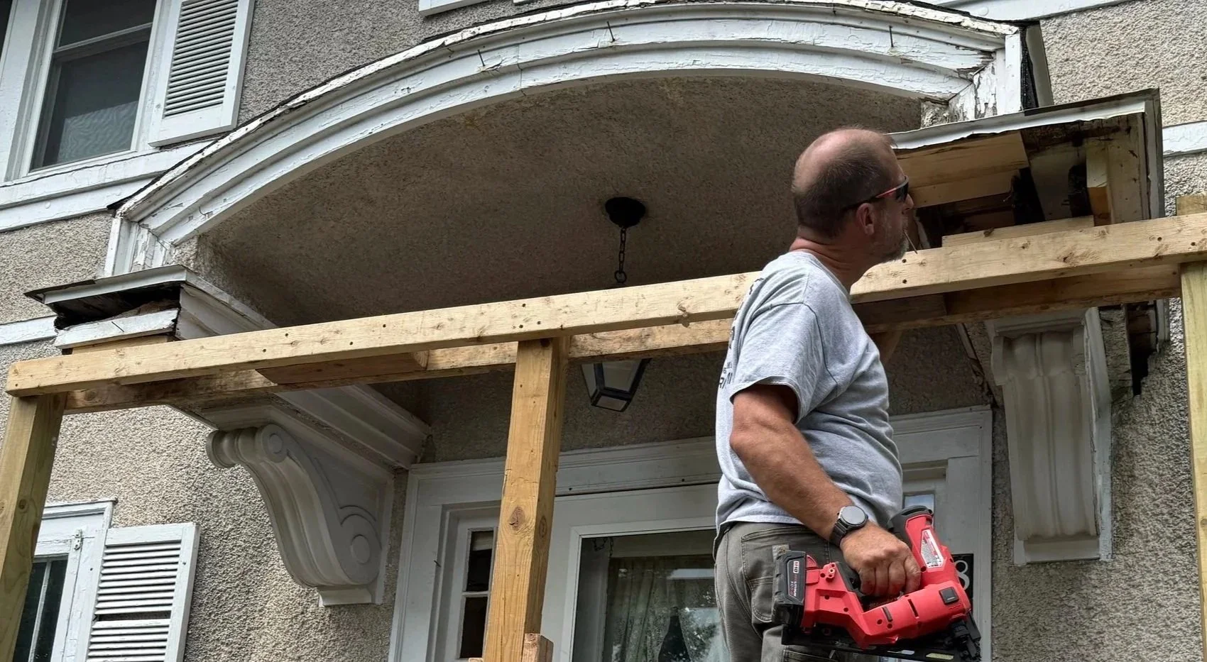 A man wearing a gray t-shirt, sunglasses, and a wristwatch stands outside a house, holding a red power drill. He is working on construction or repair around a balcony area, with wooden beams and a partially built or repaired structure.