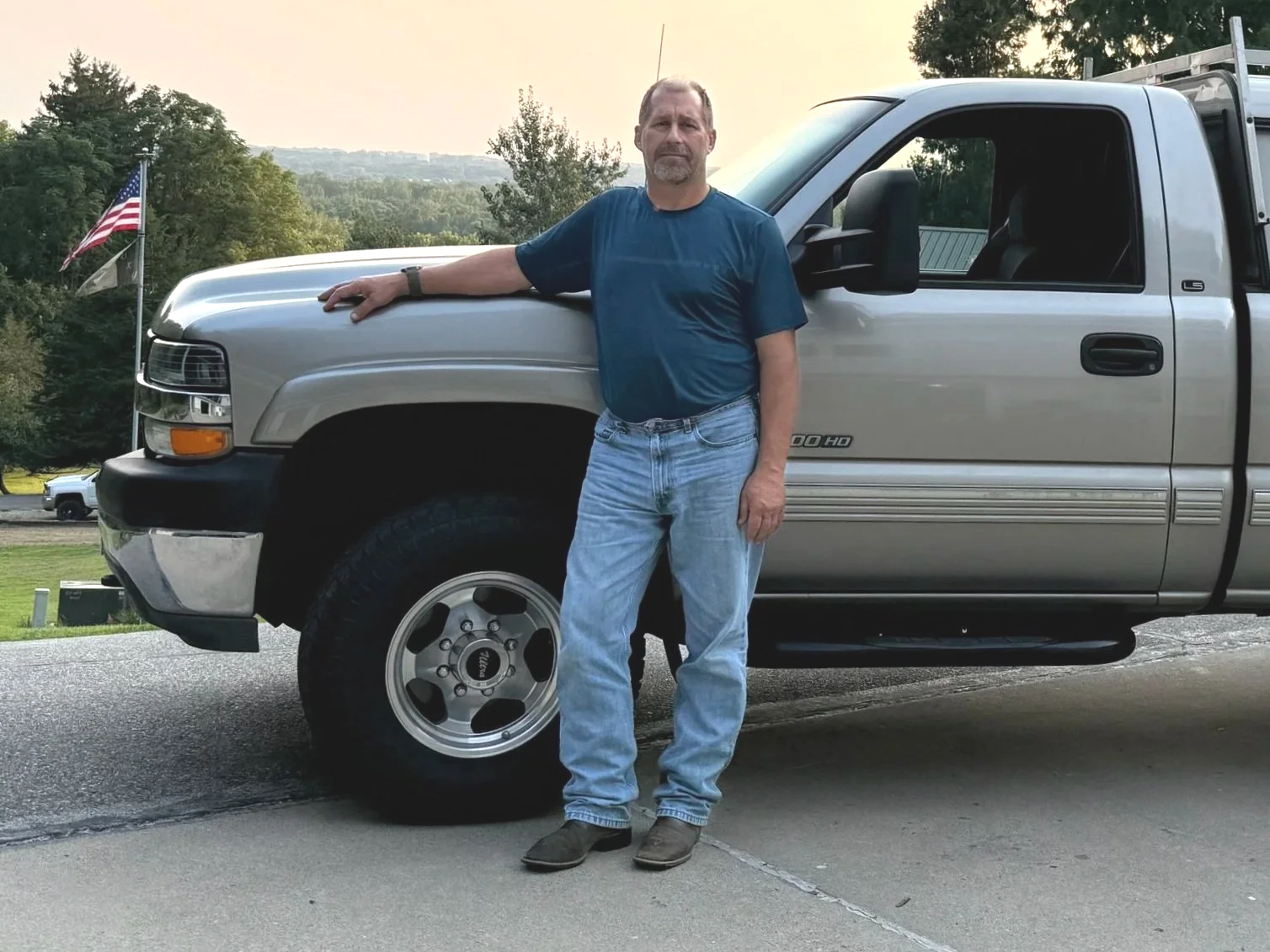 Daryl Trautmiller with DS Home Solutions standing next to his work pickup truck with his hand resting on the hood, outdoors during sunset, with trees and a flag in the background.