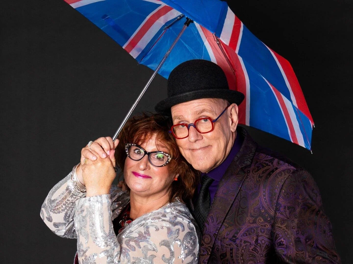 A man and woman posing under a Union Jack umbrella against a dark background. The woman has curly red hair, glasses, and is wearing a patterned jacket. The man is wearing a bowler hat, red glasses, and a patterned jacket.
