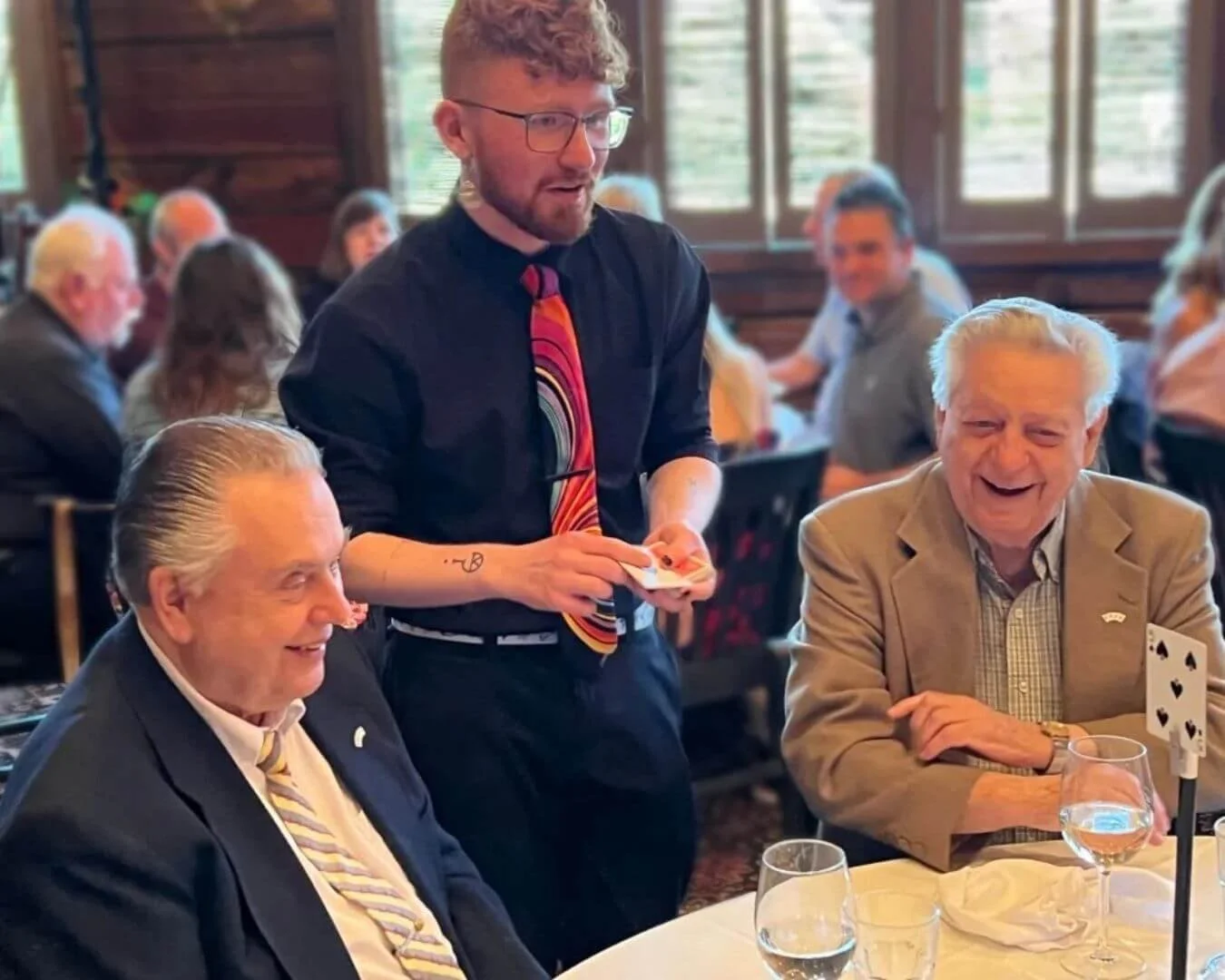 Three men sitting at a table in a restaurant, two older men laughing, and a young man standing, holding a notecard. The restaurant has wood paneled walls, large windows, and other diners in the background.