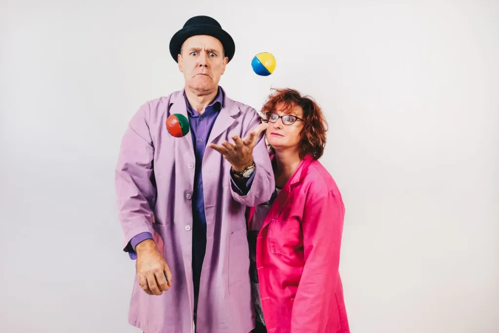 A man and a woman juggle colorful balls against a plain white background. The man is wearing a lavender coat, purple shirt, and a black hat, with a serious expression. The woman is wearing a bright pink coat and glasses, observing the balls.