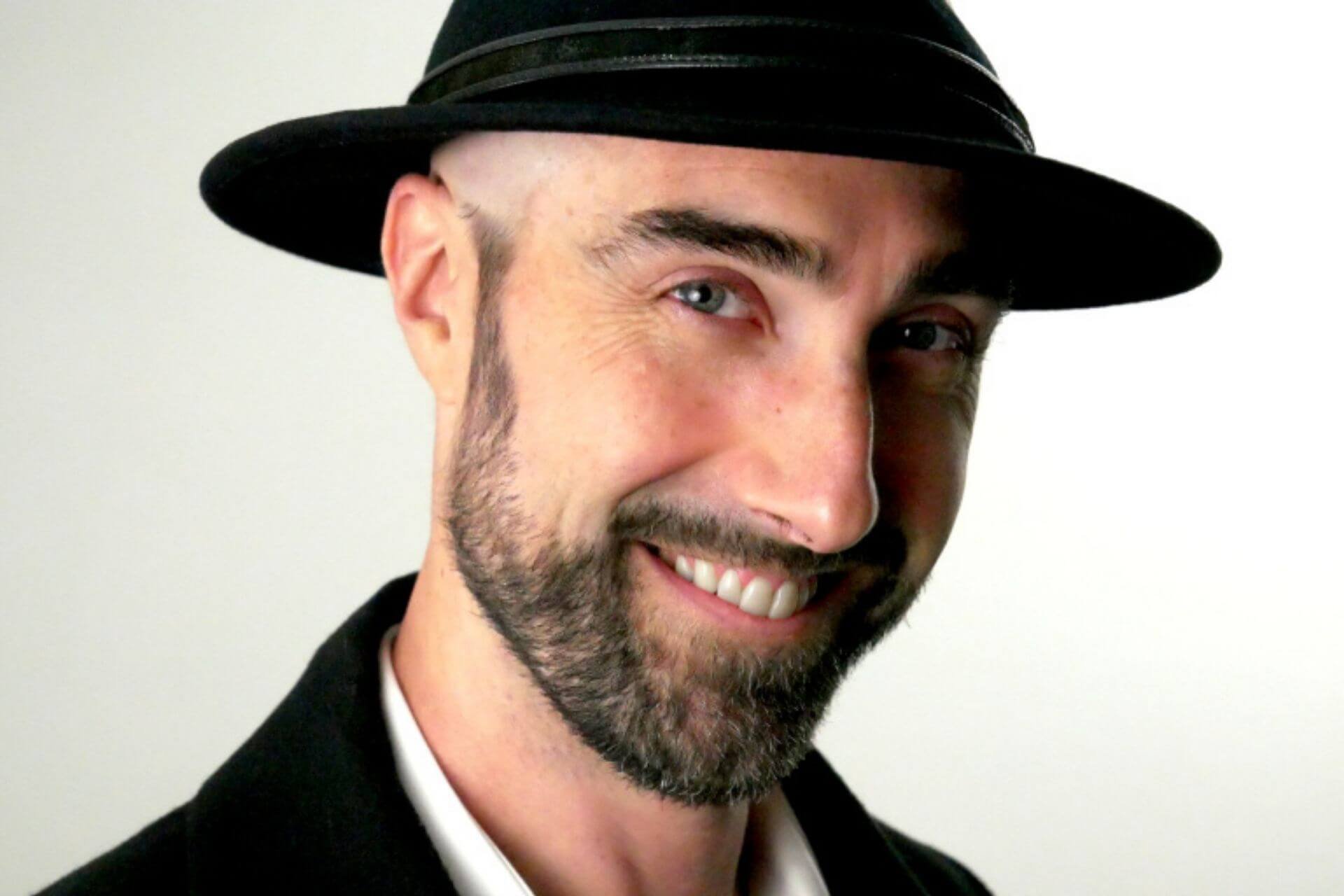 A smiling man with light skin, facial hair, blue eyes, wearing a black fedora hat with a silver band, a black jacket, and a white shirt, standing against a plain white background.