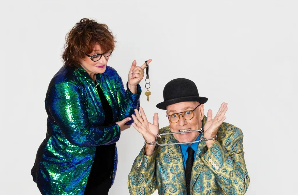 A woman holding a set of keys and a man with a clown nose and handcuffs around his neck, both wearing colorful clothing and glasses, standing against a plain white background.