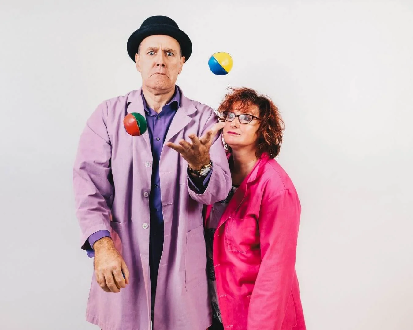 A man in a purple coat and black hat juggling colorful balls, with a woman in a pink coat watching.