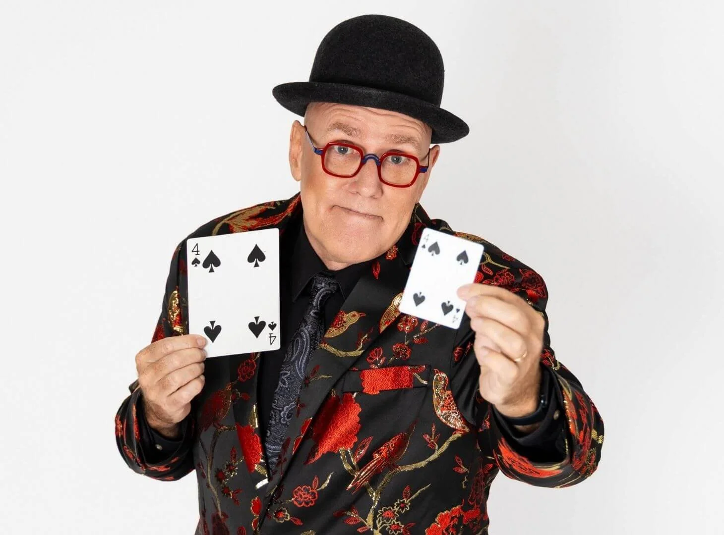 Man wearing a black bowler hat and colorful patterned blazer showing two playing cards, the four of spades and the two of spades, against a plain white background.