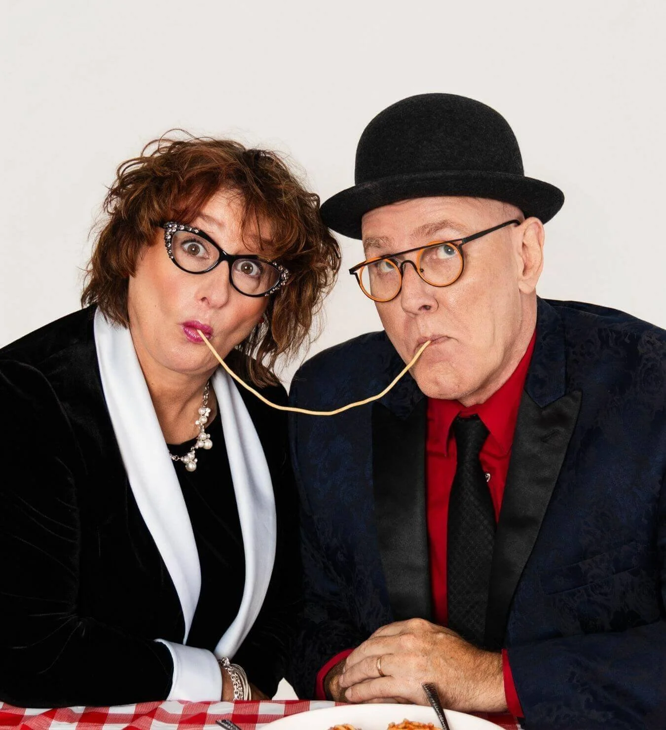 A man and woman dressed in formal attire with spaghetti in their mouths, connected by the spaghetti, sitting at a table with a red and white checkered tablecloth.