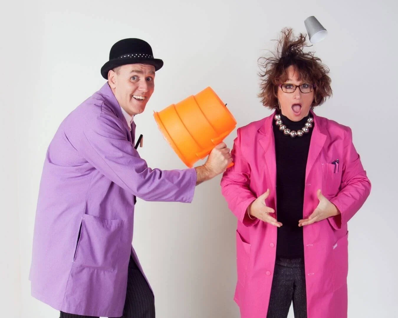 A man and a woman dressed in colorful coats, with the man holding an orange lamp and the woman showing surprise, against a plain background.