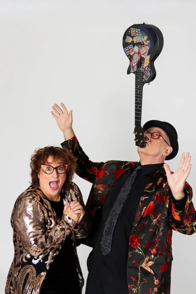 A woman and a man with glasses and colorful clothing, excited and smiling, holding a guitar-shaped case filled with British flags and accessories, with the man balancing the case on his lips.