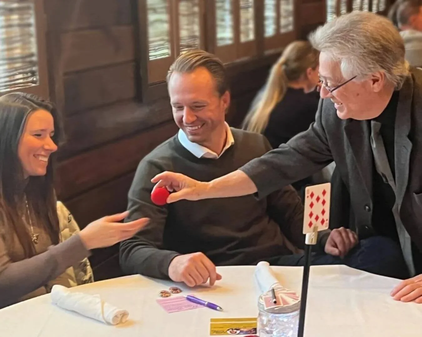 A group of people sitting at a table in a restaurant, smiling and enjoying a magic card trick performed by an older man with gray hair and glasses. The older man is holding a red ball, and there is a playing card on a stand on the table.