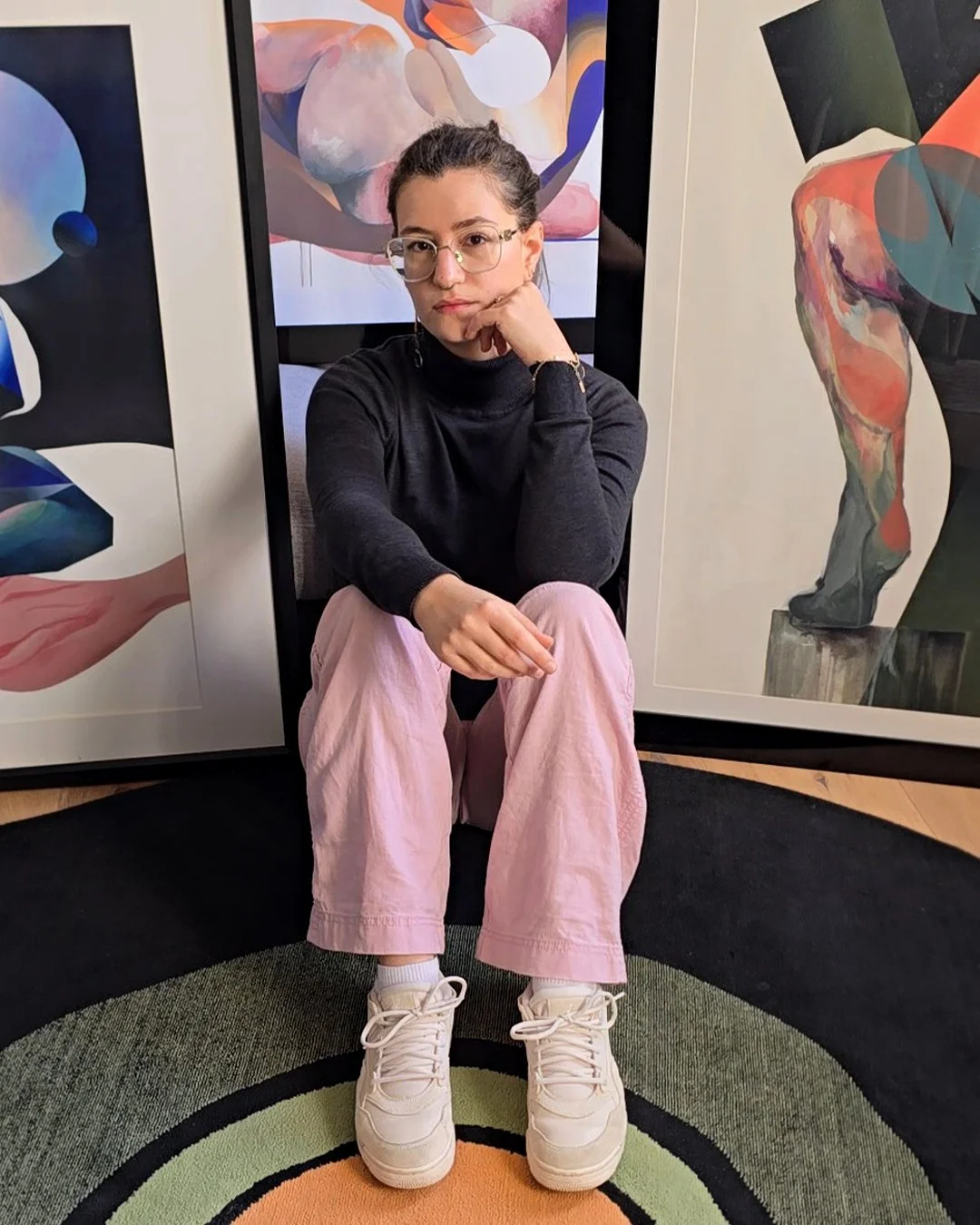 Image de portrait de l'artiste devant ses œuvres d'art, portant un pull noir, un pantalon rose pâle, des chaussures blanches et des lunettes.